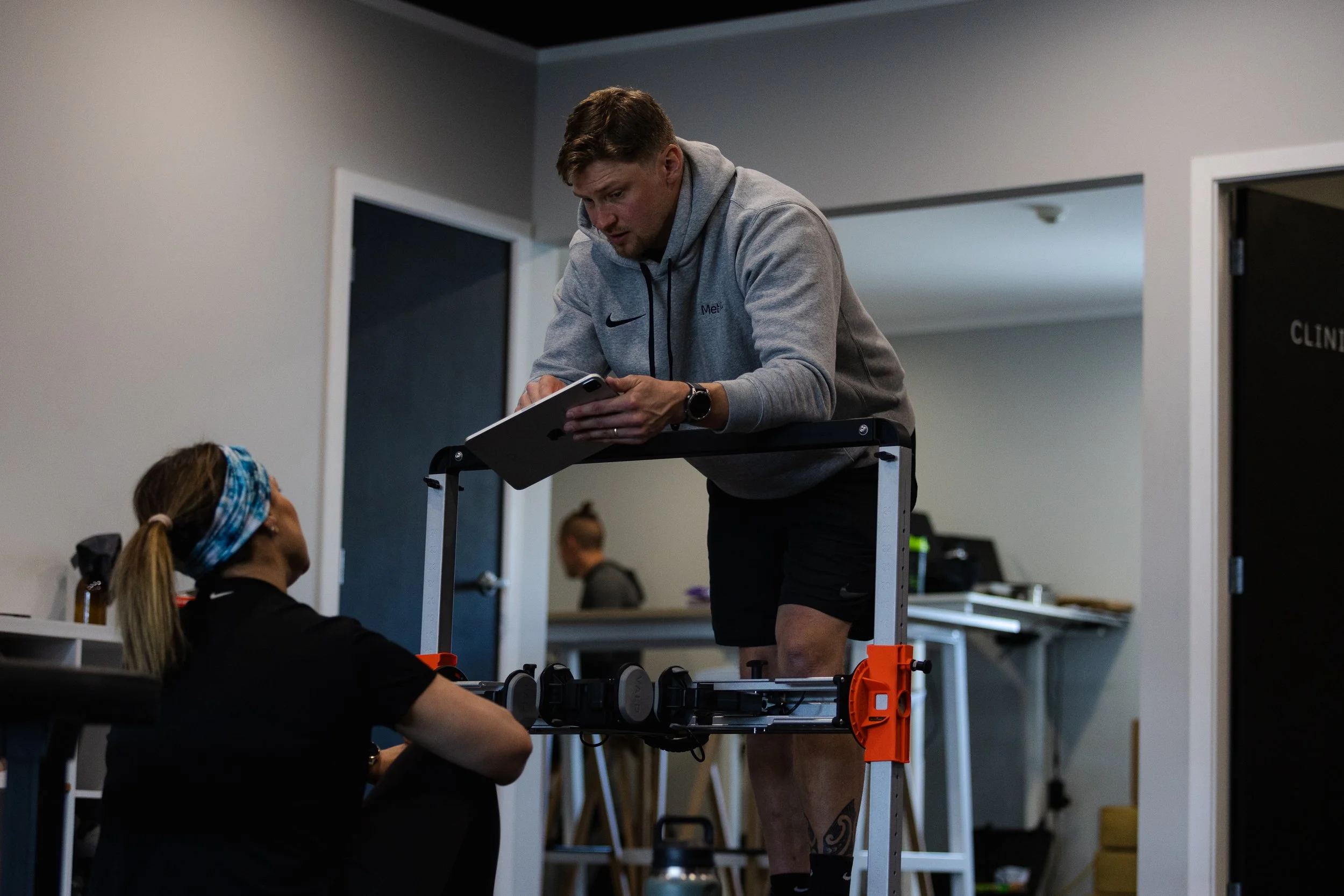 A man with short hair in a gray hoodie and black shorts is leaning over a table, looking at a tablet. A woman with a ponytail and headband is seated at the table, also looking at the tablet. The scene takes place indoors, possibly at a gym or physical therapy clinic.