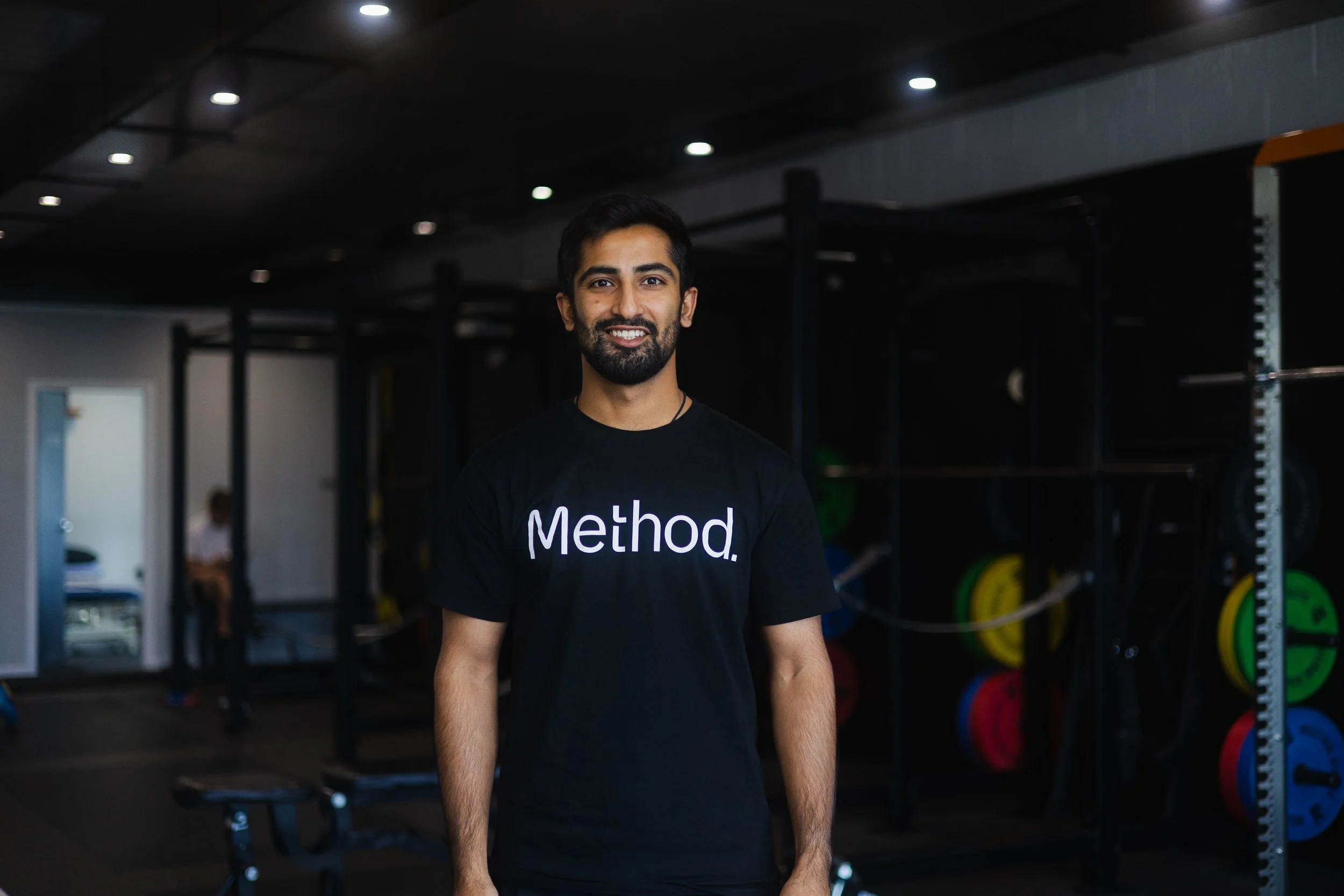 A smiling young man with a beard and dark hair stands in a gym, wearing a black T-shirt with the word 'Method' printed on it. The gym is equipped with weightlifting equipment and colorful weight plates in the background.