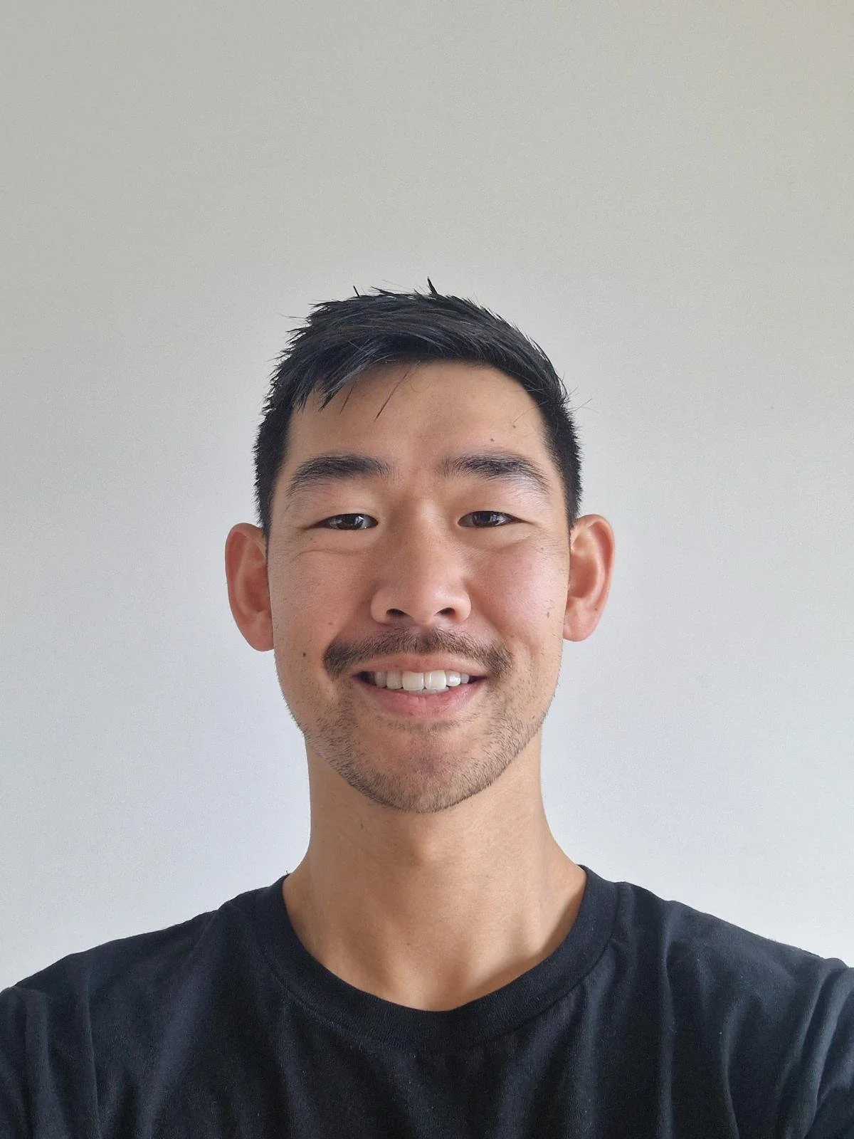 A young man with short black hair, a slight beard, and a mustache, smiling at the camera against a plain white wall. He is wearing a black T-shirt.