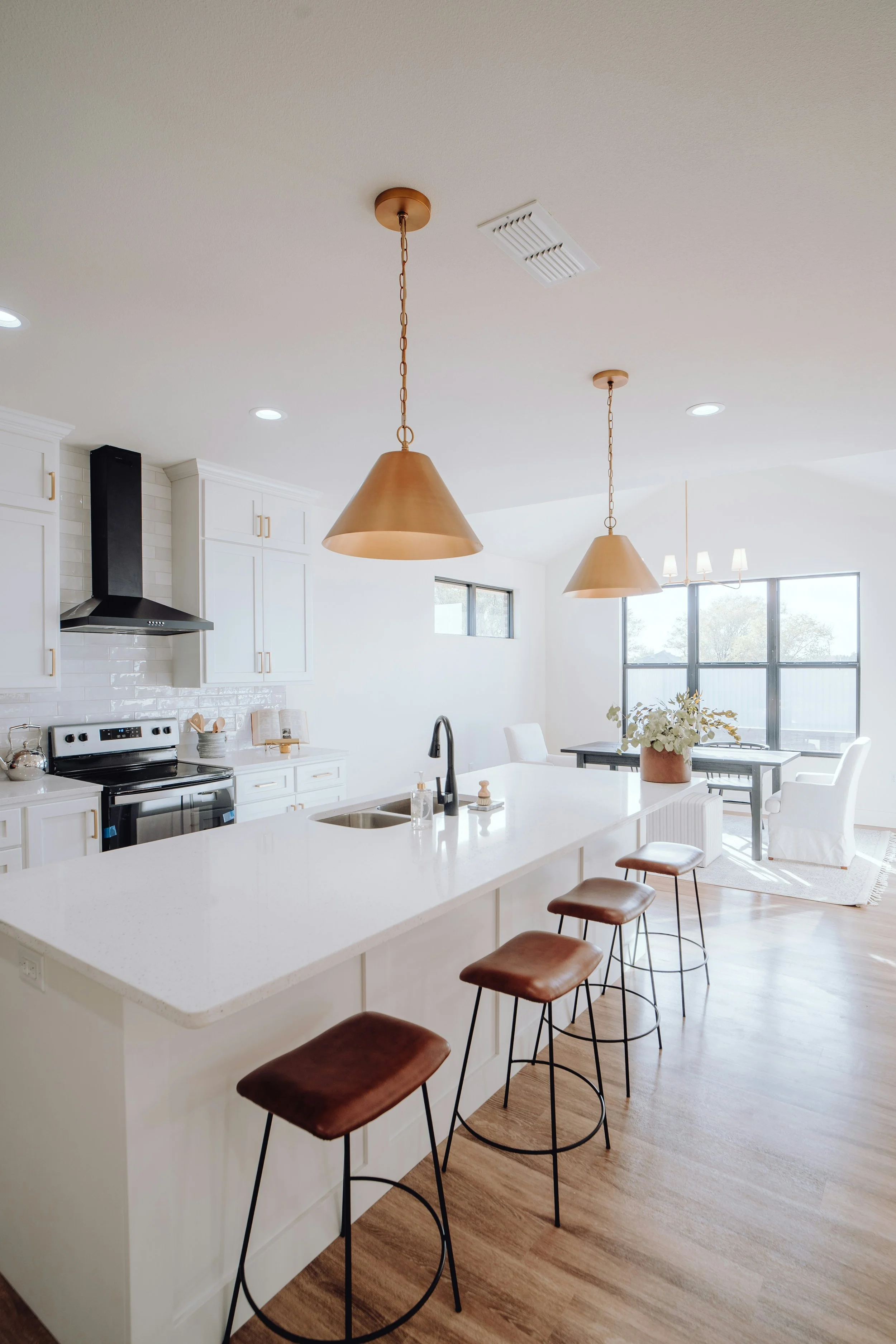 Bright, modern kitchen and dining area with white cabinets, a white island with a sink, black faucet, and brown bar stools. Two large brass pendant lights hang over the island, and natural light streams in through large windows. There are also ceiling lights and a small floral arrangement on the dining table.