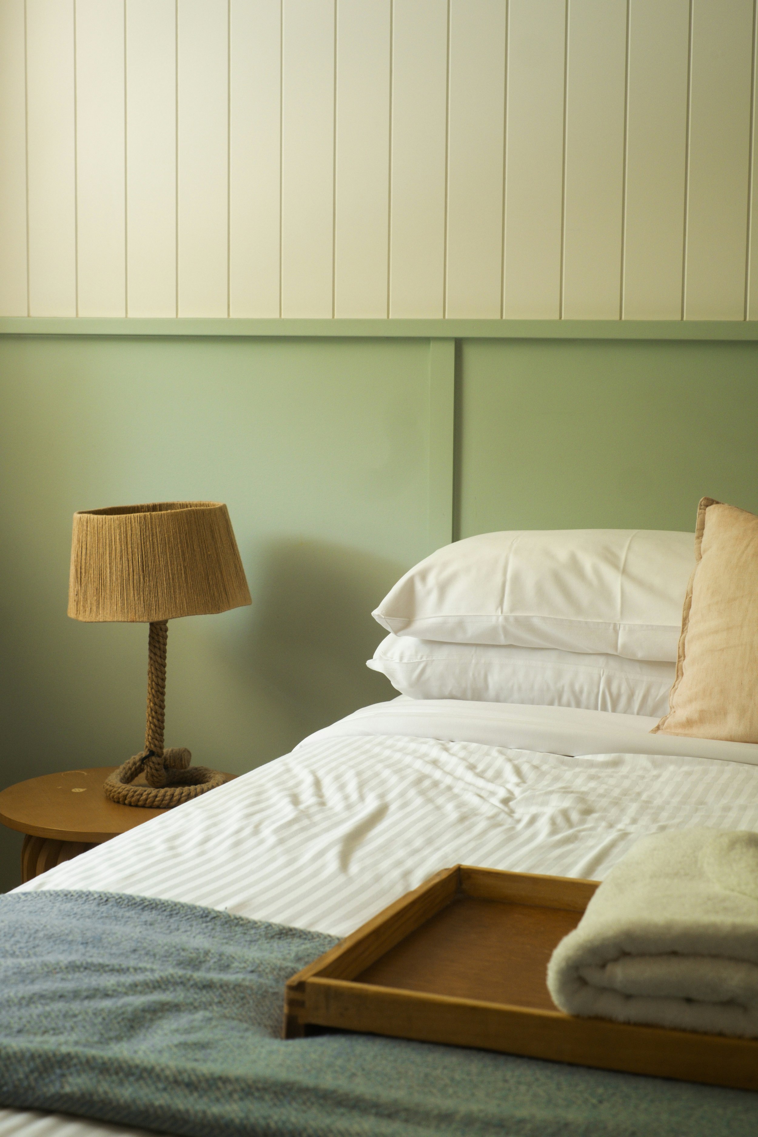 A neatly made bed with white sheets and two white pillows, a beige pillow, and a bed tray with two rolled towels, in a room with pale green walls, a wooden side table, a braided lamp, and a wooden tray.