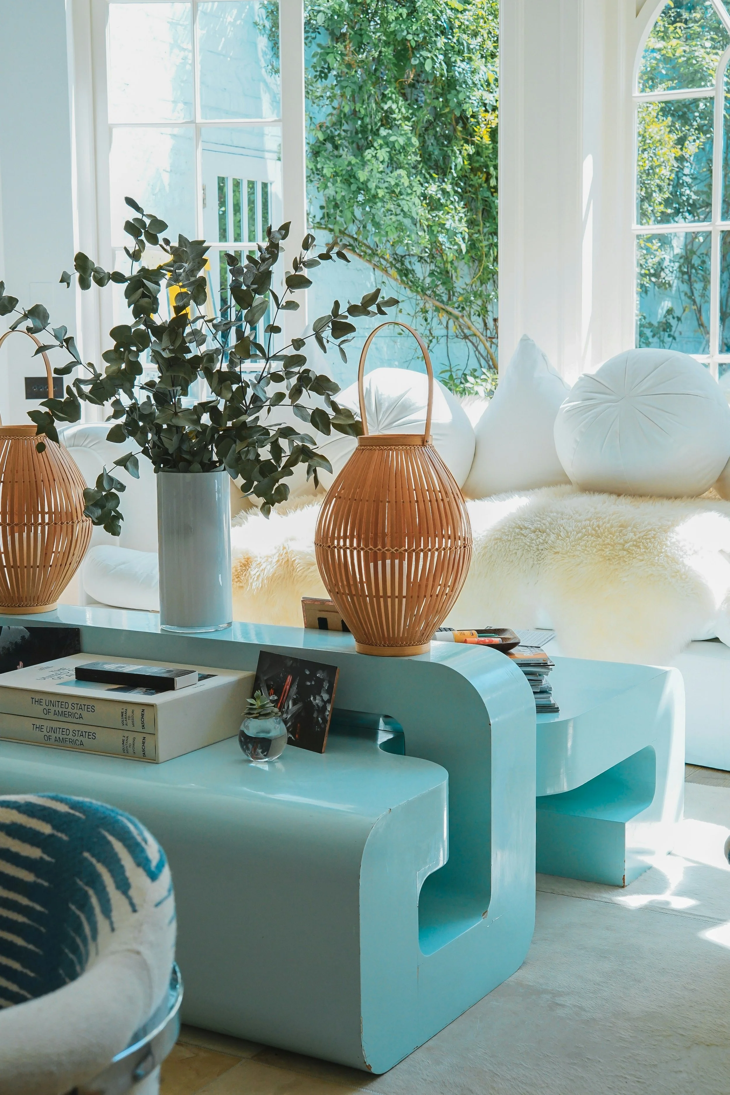 A cozy living room corner with a large window overlooking greenery. The space features white cushions, a plush cream rug, a large leafy plant in a white vase, and two wicker lanterns on a white curved coffee table. Books and magazines are also on the table.