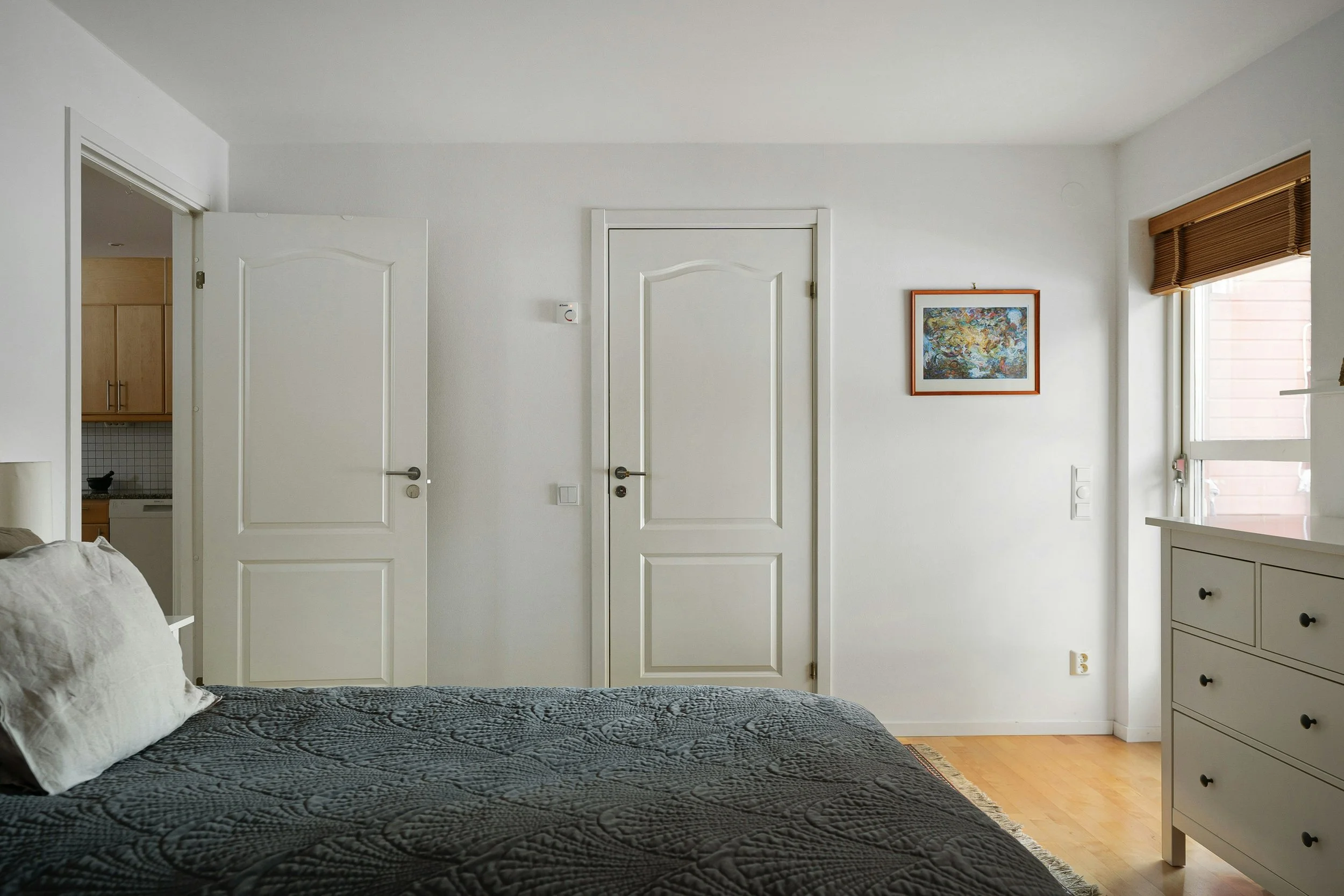 Interior view of a bedroom with a bed in the foreground, white walls, a framed colorful artwork on the wall, a window with brown blinds, and two closed white doors.