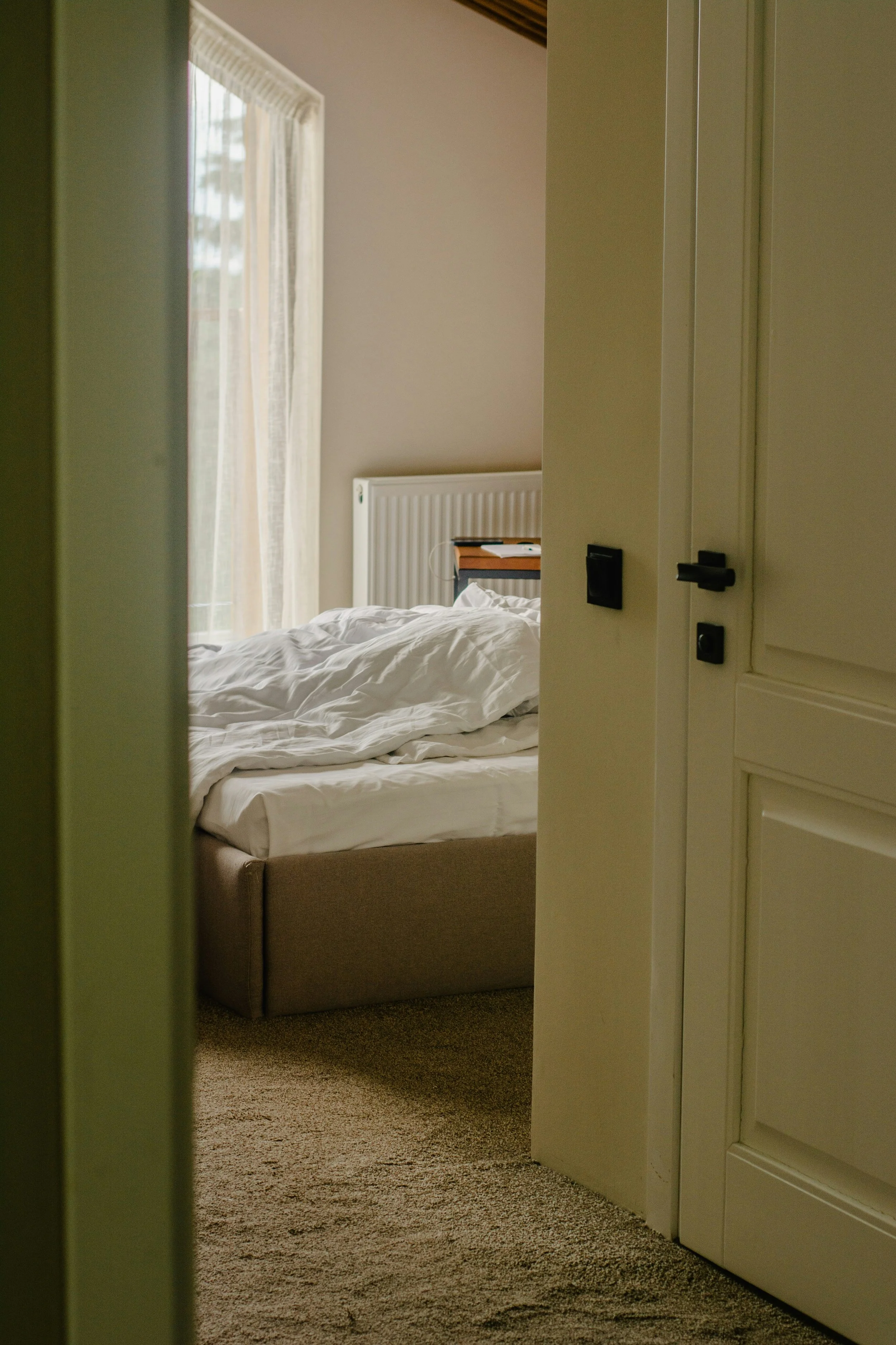 A view of a bedroom through a partially open door. The bed, with white sheets, is unmade and sits against a wall beneath a window with curtains. A radiator and a small bedside table with some items are visible in the room.