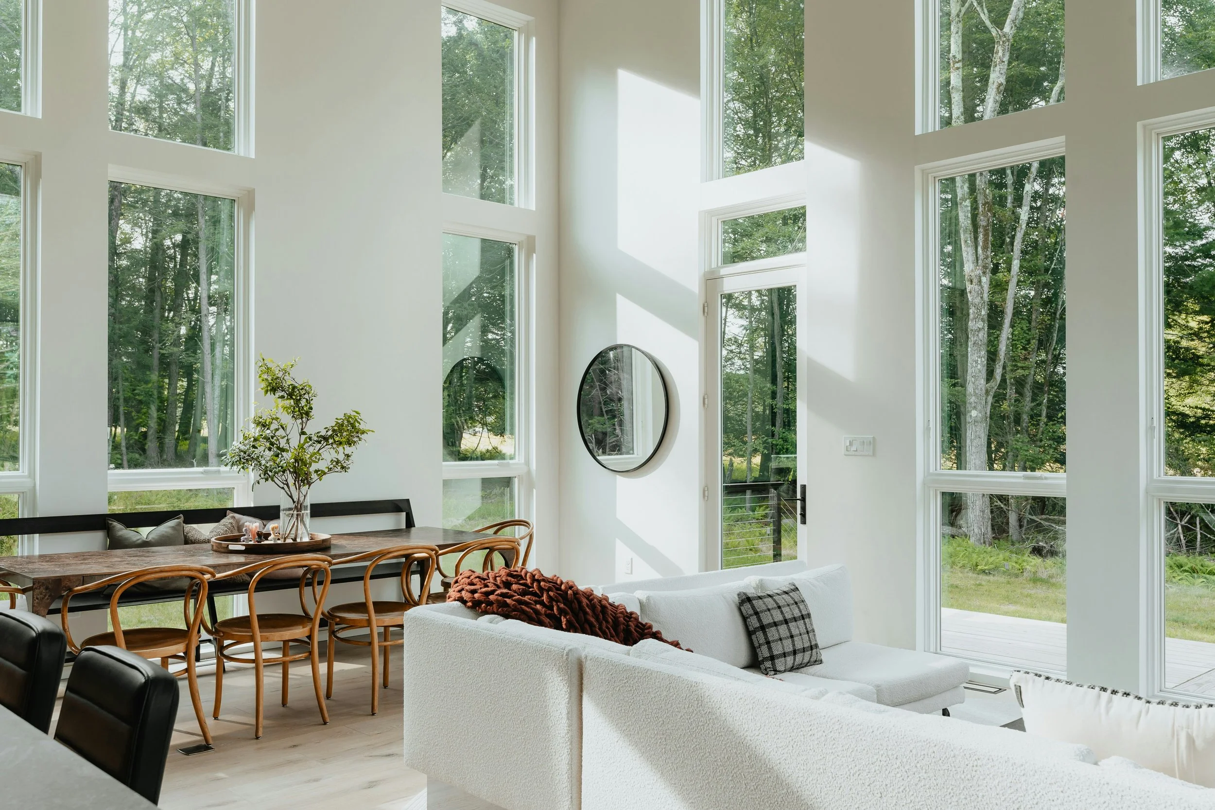 A bright living room with large, floor-to-ceiling windows showing a view of trees outside. The room features a white sofa with a throw blanket, a round mirror on the wall, and a wooden dining table with chairs.