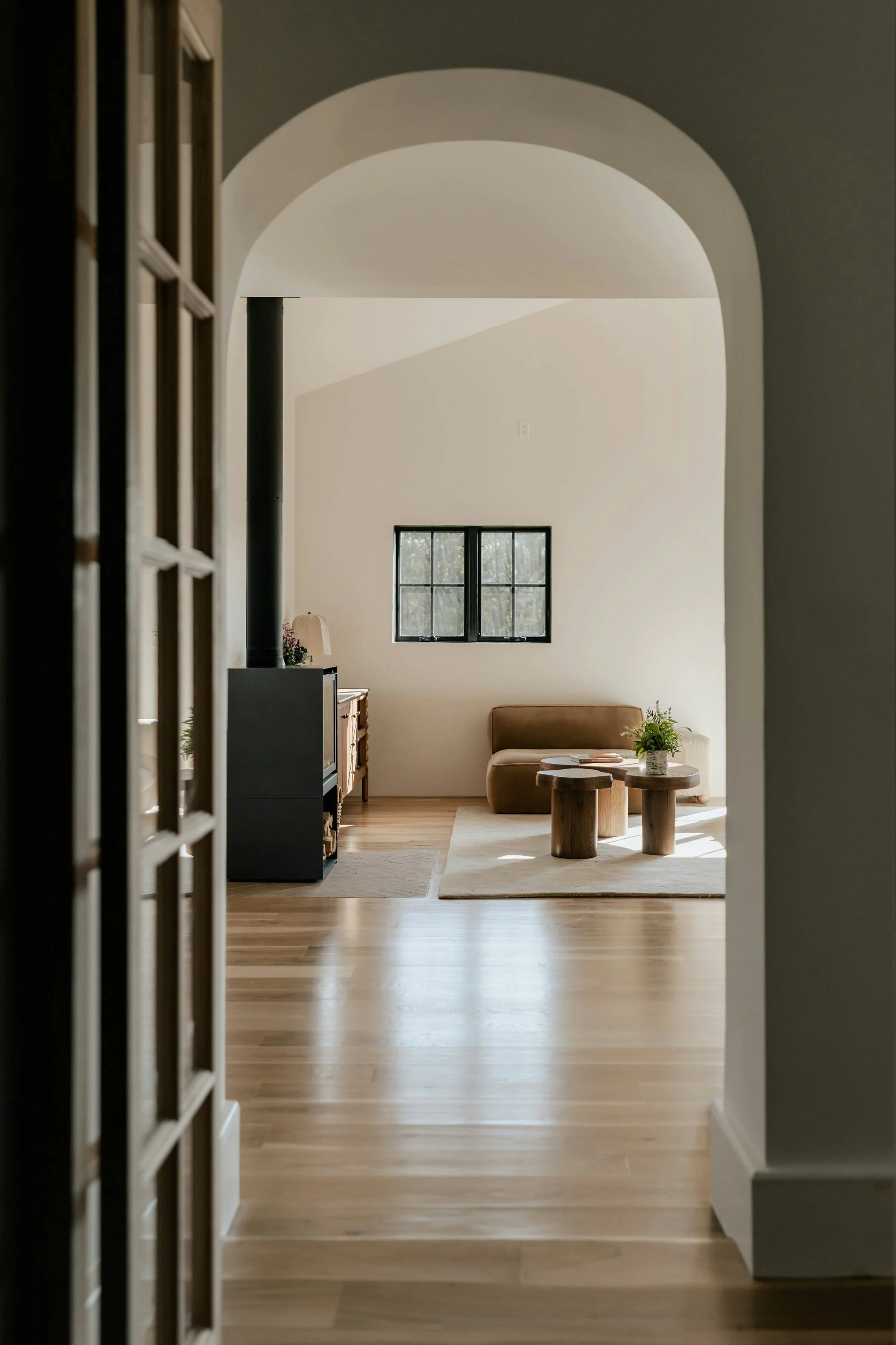 View through an arched doorway into a bright living room with a window, a sofa, and a wooden coffee table with a plant on it.