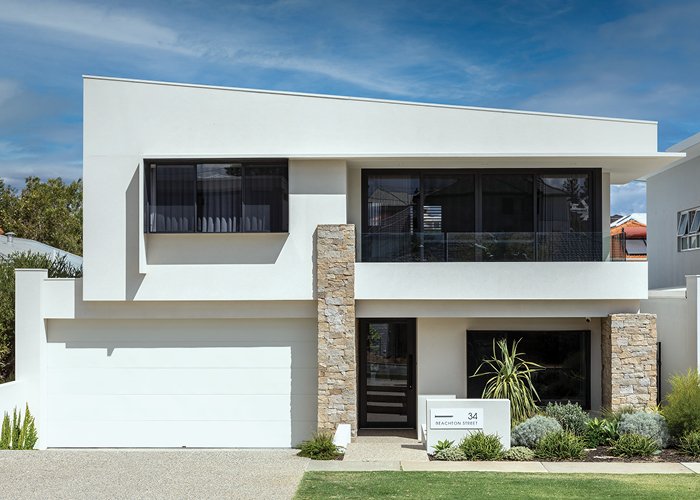 Modern two-story white house with stone accents, large windows, and a garage. Landscaping includes shrubs and grass, with a street number sign near the entrance.