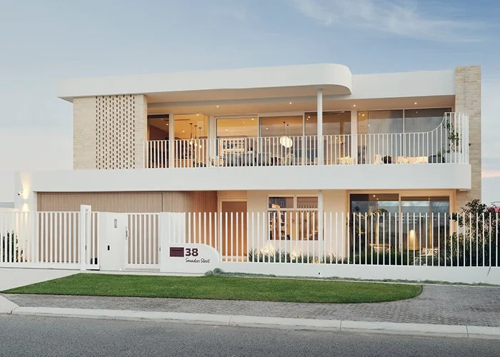 Modern two-story house with white exterior, large glass windows, and a balcony with a white railing. The house has a front yard with grass and is enclosed by a white fence. The house number is 138.