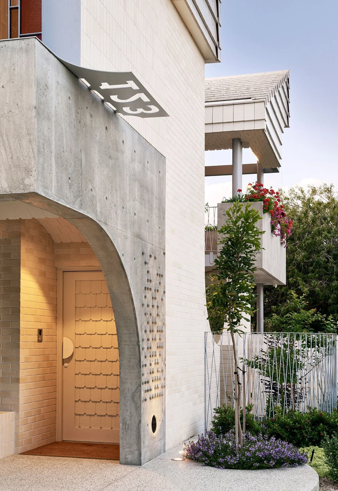 Modern house with a concrete and brick facade, arched entrance, and a balcony with pink and red flowers, a small tree, and a white metal fence in front.
