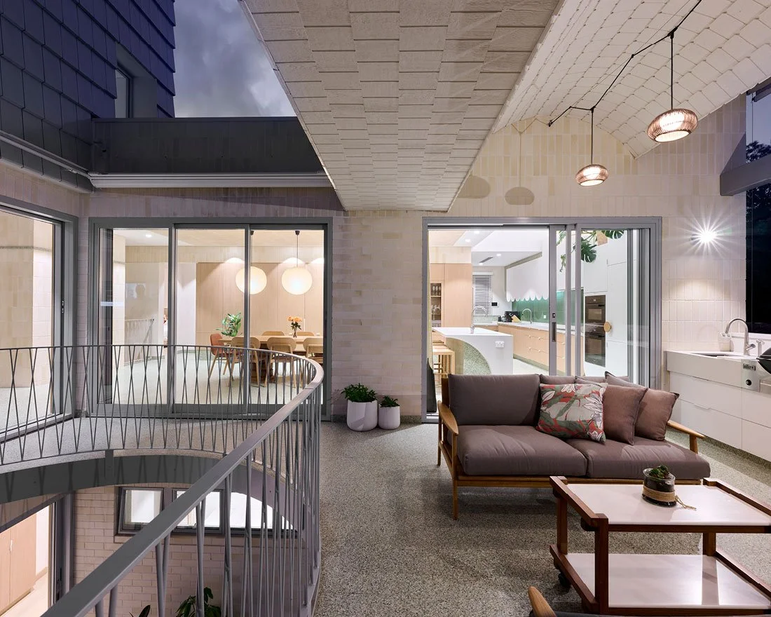 Modern living room with large windows, a gray sofa with pillows, a wooden coffee table, the kitchen visible through sliding glass doors, and a balcony with a curved railing.