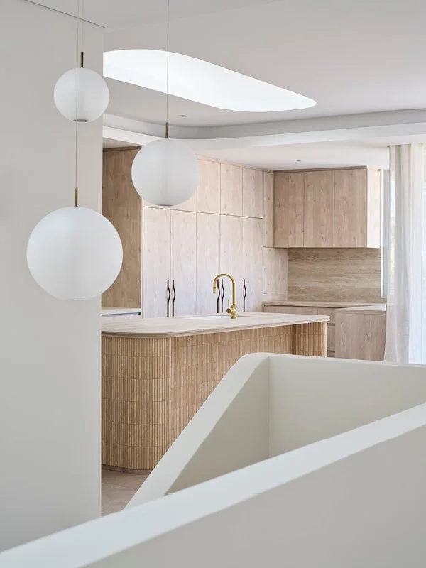 Modern kitchen with light wood cabinets, white countertops, a curved island with vertical wood paneling, gold faucet, and three hanging white pendant lights.