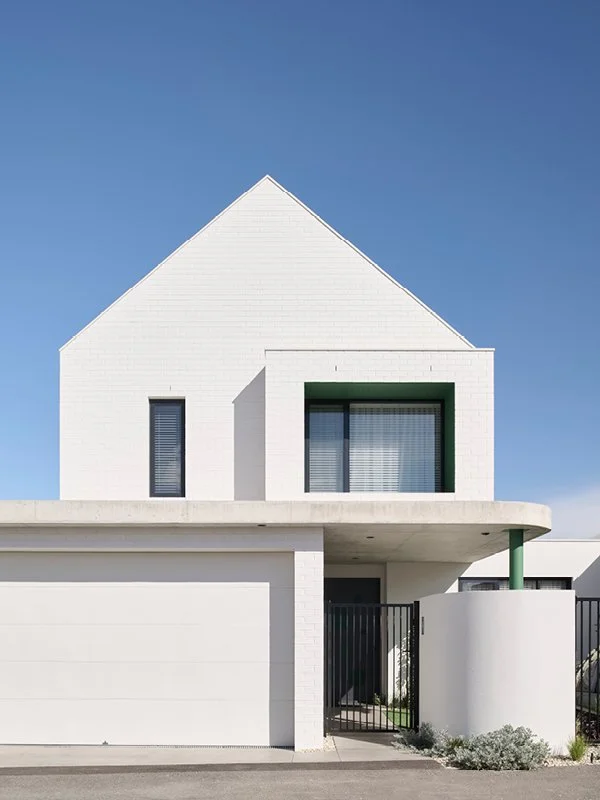 Modern white house with a peaked roof, large glass window, and black gate, set against a blue sky.