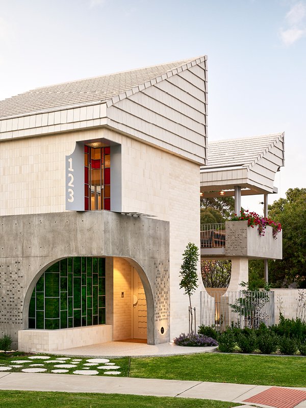 Modern residential house with a concrete archway entrance, upper floors featuring large windows, and a balcony with potted plants, surrounded by landscaped garden and greenery.