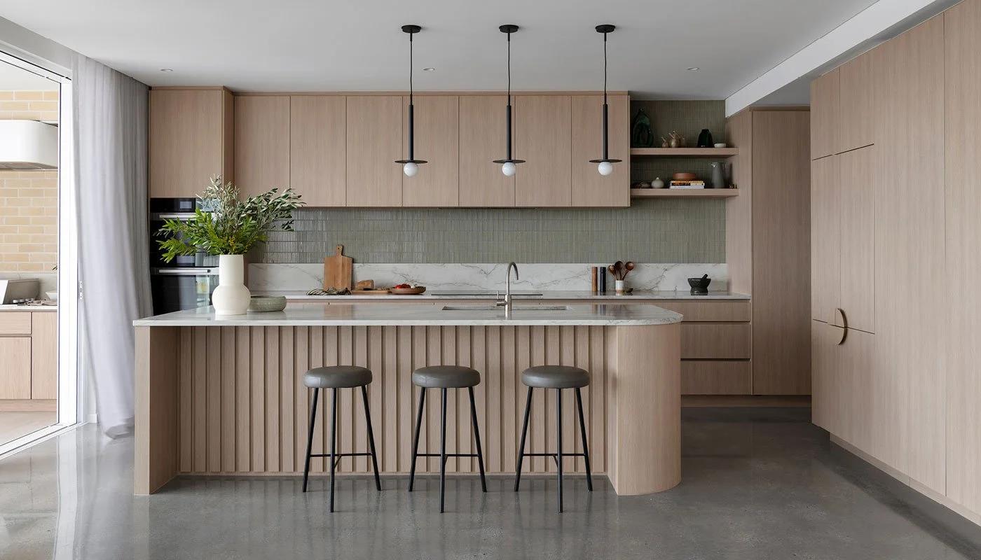 Modern kitchen with light wood cabinets, marble countertop and backsplash, three black pendant lights, and three gray bar stools at the breakfast bar