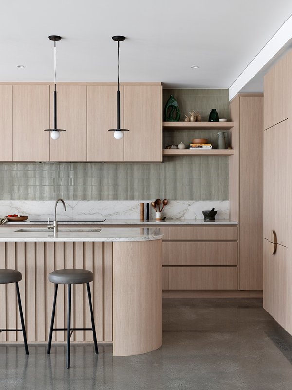 Modern kitchen with light wood cabinets, a marble island with two barstools, green tile backsplash, open shelves with decorative items, and pendant lights.