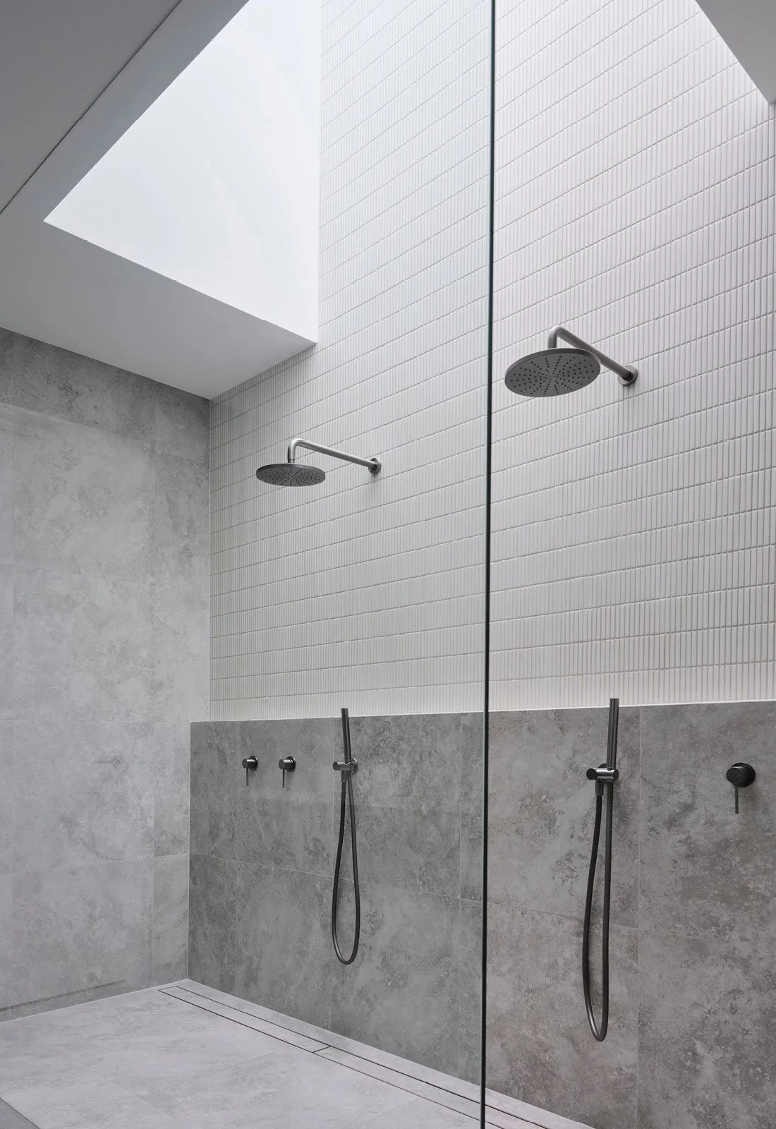 Modern shower area with two rain showerheads and handheld showers, white tiled wall, gray concrete lower wall and floor, and a skylight above.
