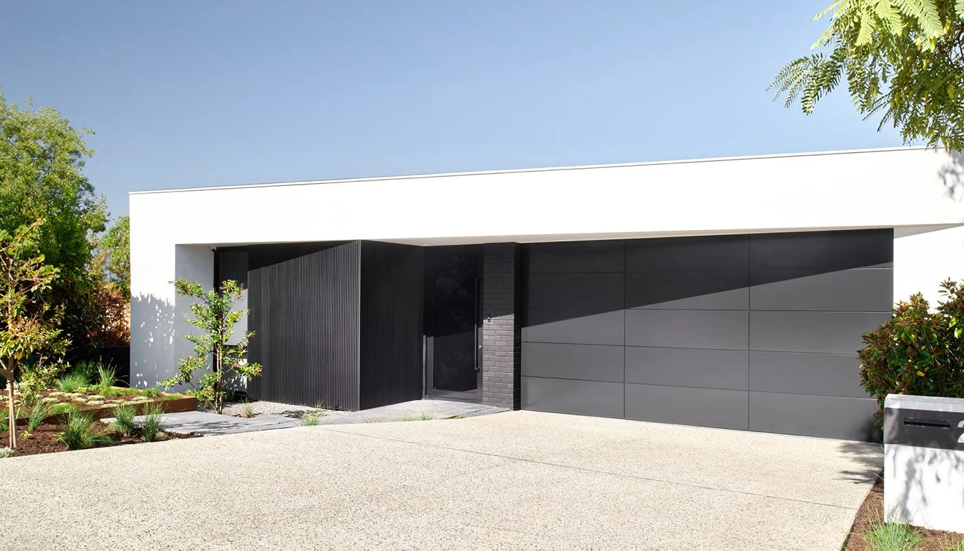 Modern house with white, black, and gray facade, attached garage, and landscaped front yard with small trees and bushes.
