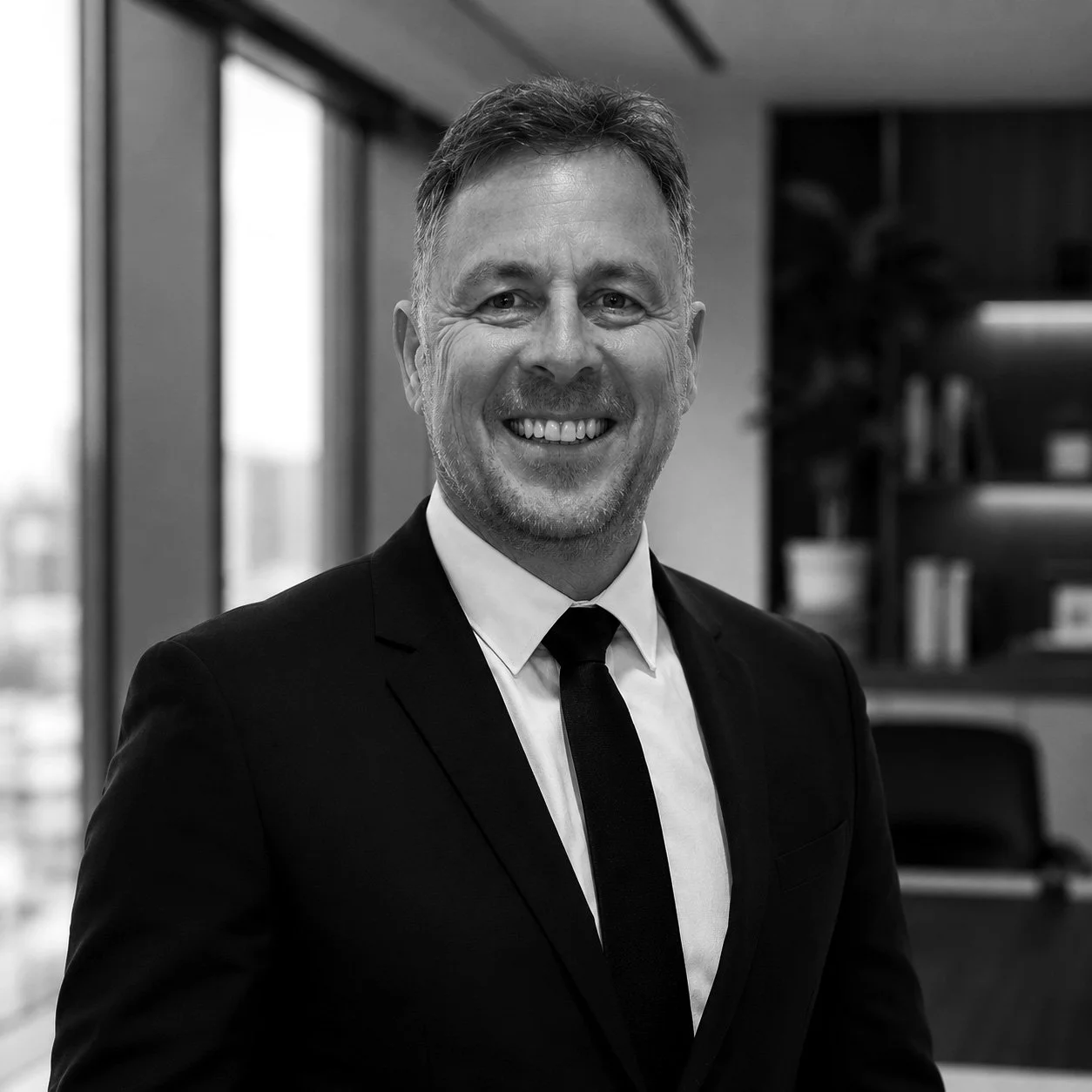 A smiling man in a suit and tie posing in an office with large windows and bookshelves in the background.