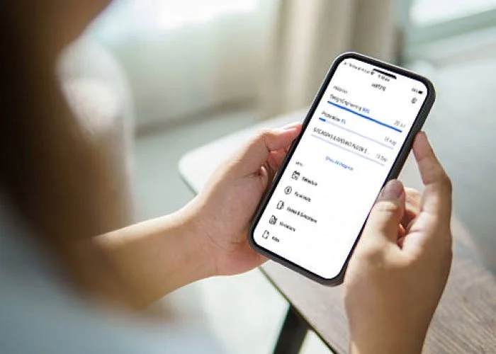 Close-up of a person holding a smartphone displaying a financial app, sitting at a table with a window in the background.