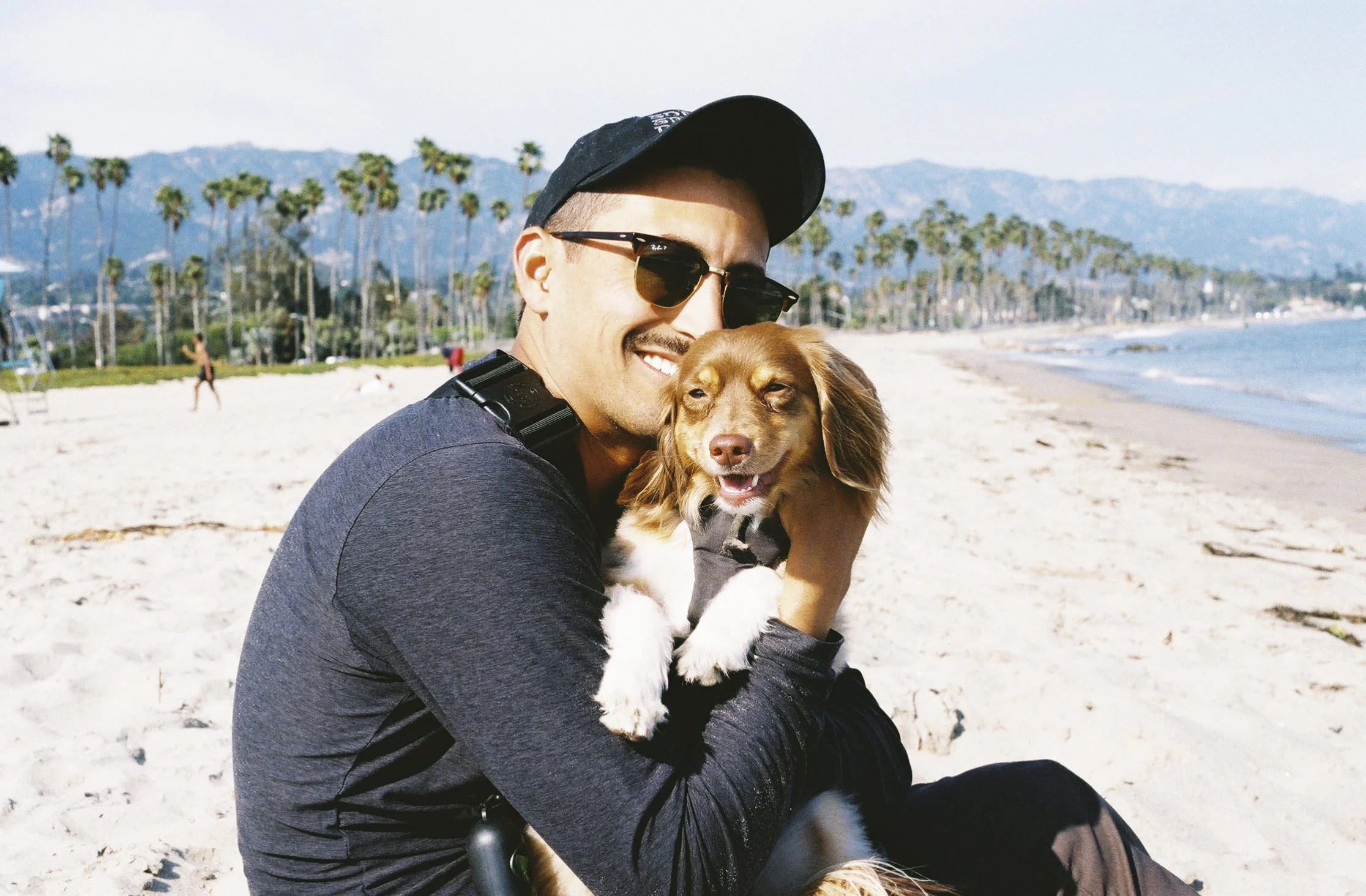 A man wearing sunglasses and a cap holding a brown and white dog at the beach with palm trees and mountains in the background.