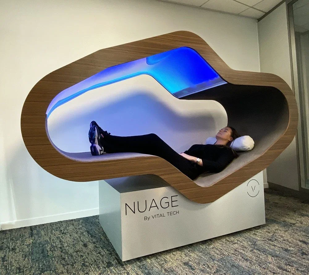 A woman lying inside a futuristic, capsule-like pod with wooden exterior and illuminated blue interior, labeled 'NUAGE By VITAL TECH'.