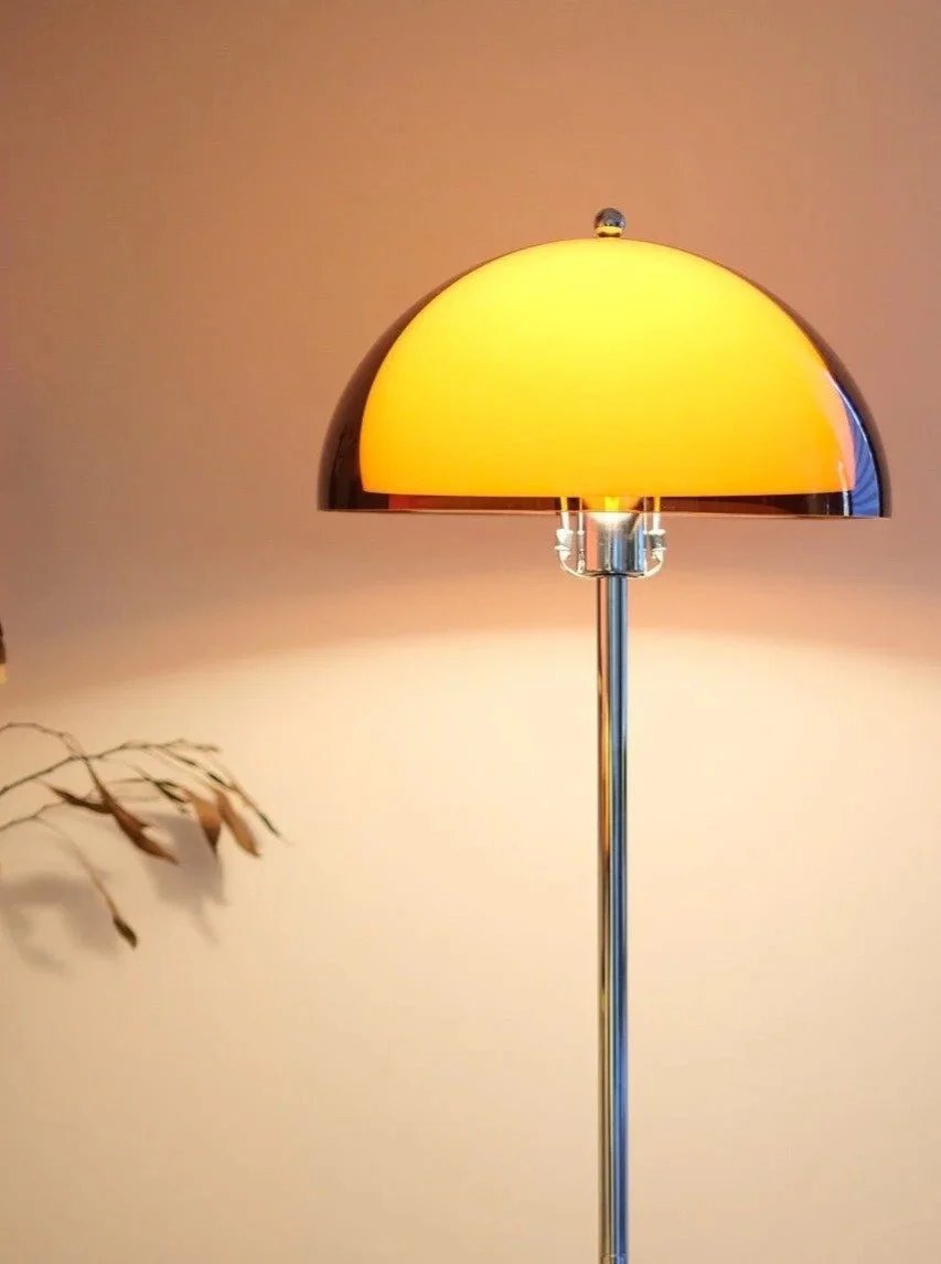 A floor lamp with a yellow lampshade and a metallic stand, standing next to a beige wall with a plant shadow nearby.