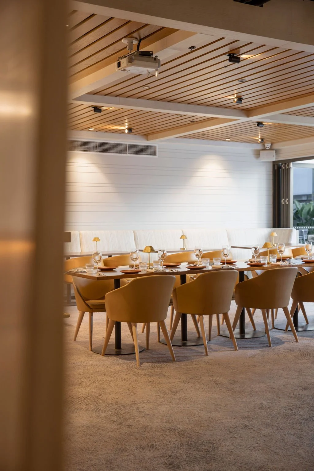 A modern restaurant dining area with a long table set with plates, glasses, and small lamps, surrounded by beige chairs, with a white paneled wall and large window in the background.