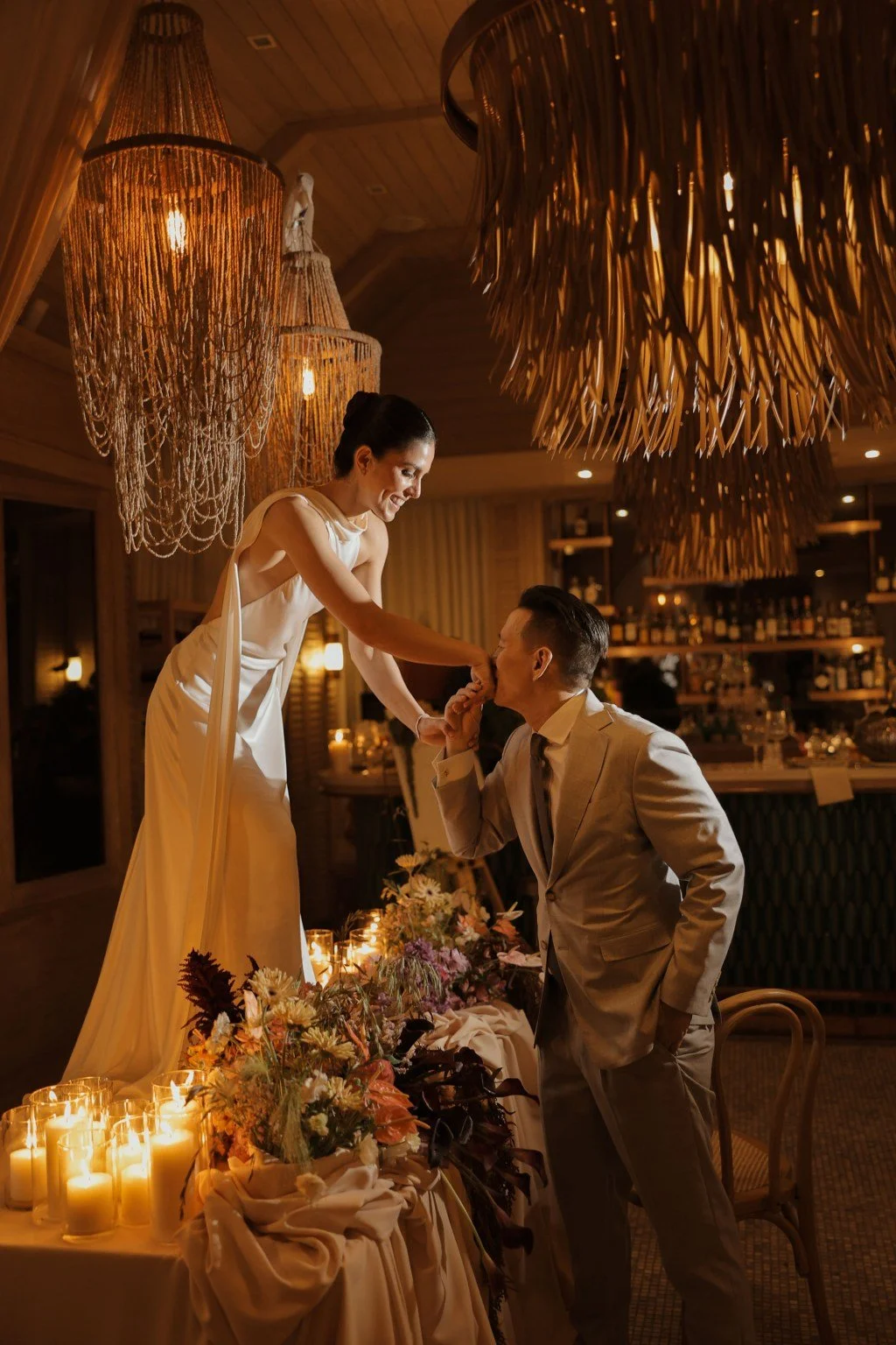 A woman in a white dress leaning over a table, holding a man's chin, kissing his fingers in a warmly lit venue with candles, flowers, and chandeliers.