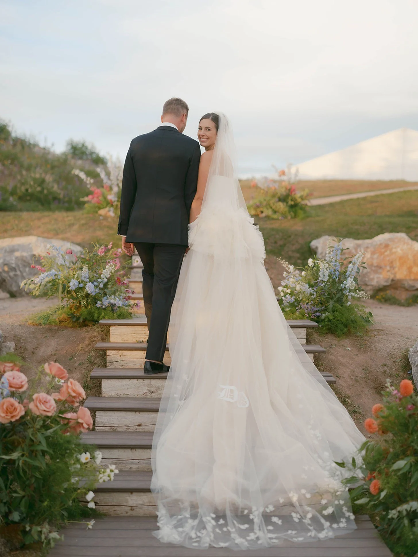 A bride and groom standing on outdoor wooden steps adorned with flowers, facing away but smiling at each other. The woman is wearing a long, flowing white wedding gown with a veil, and the man is in a black tuxedo. The background features a cloudy sky and a garden landscape with rocks and flowers.