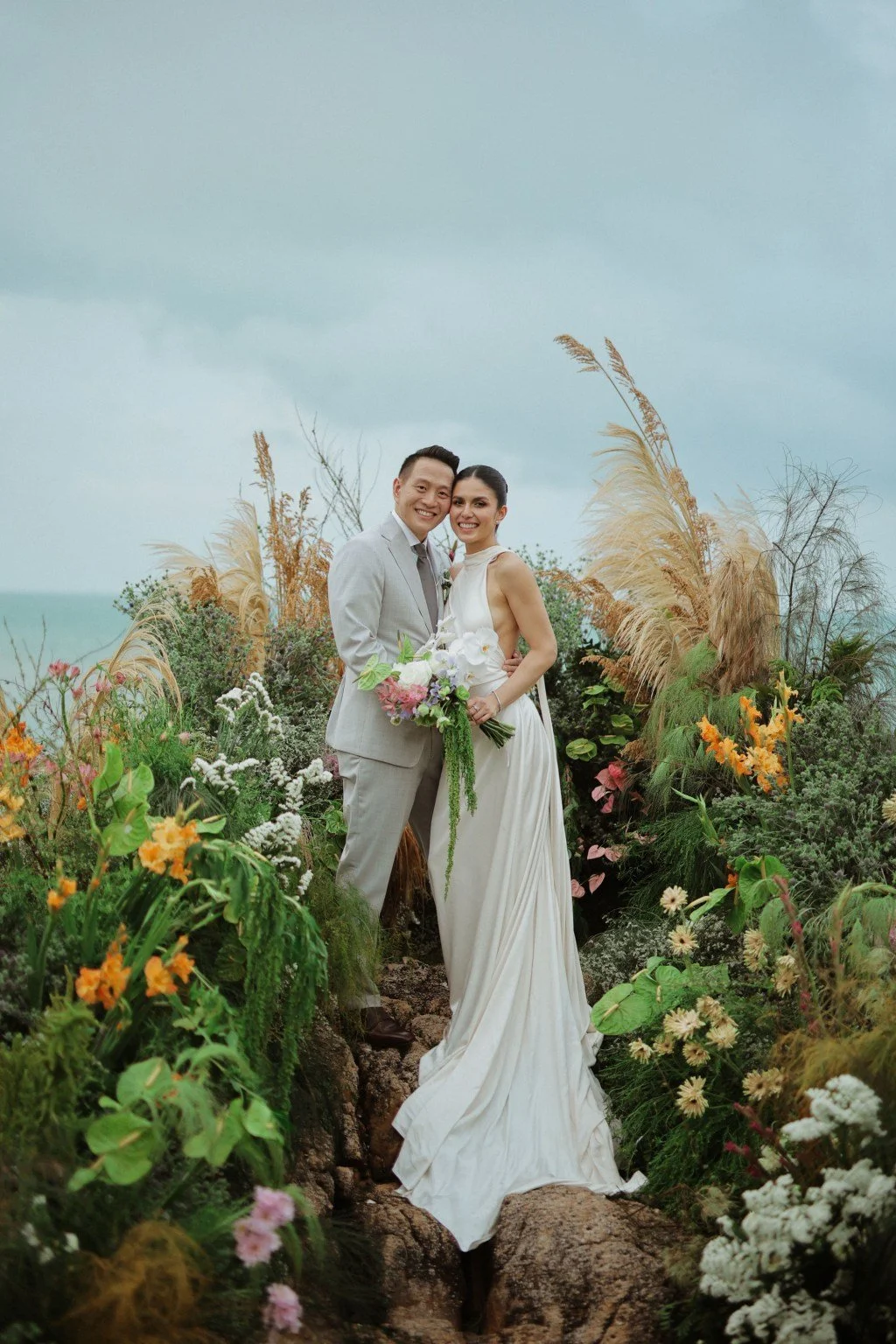 A newlywed couple in wedding attire standing on rocky ground surrounded by colorful flowers and tall grasses with cloudy sky in the background.
