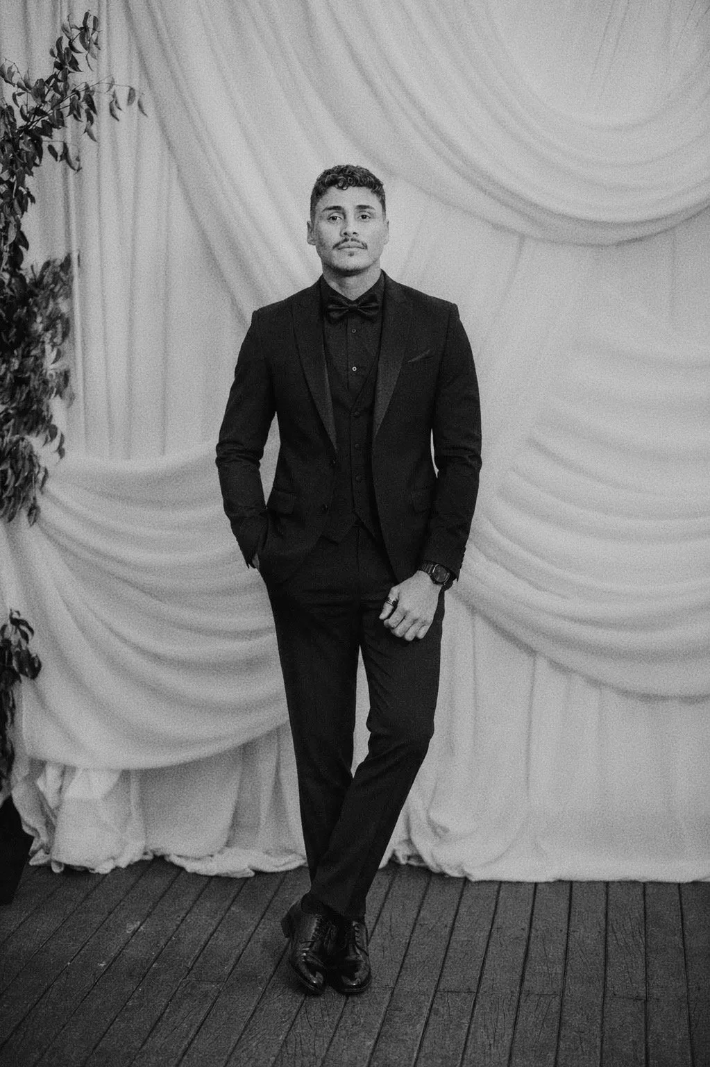 A man wearing a tuxedo standing in front of a draped curtain backdrop, with one hand in his pocket and the other resting by his side. He is looking at the camera with a confident expression.