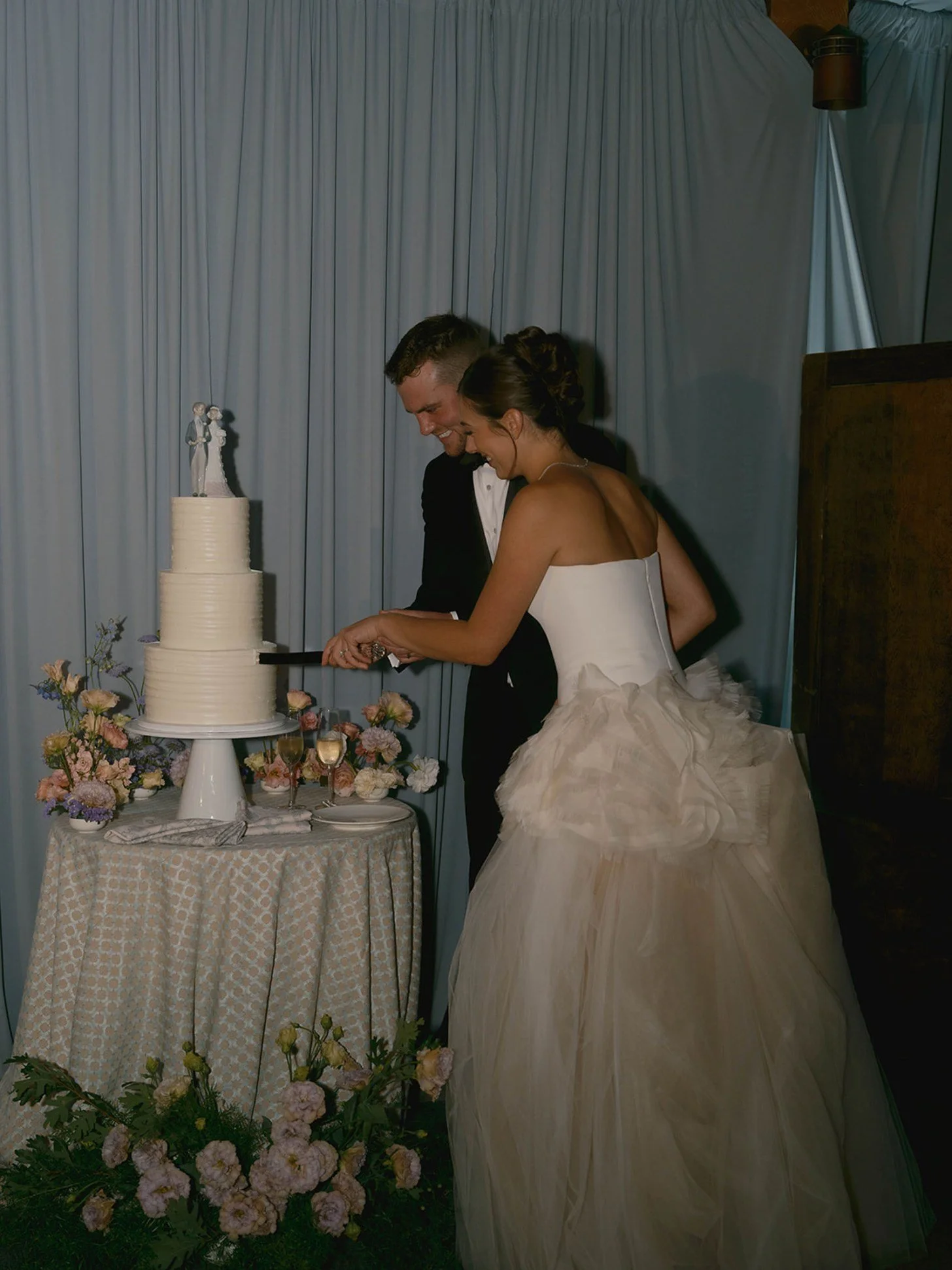 A bride and groom cut into a wedding cake at their wedding reception, surrounded by flowers.