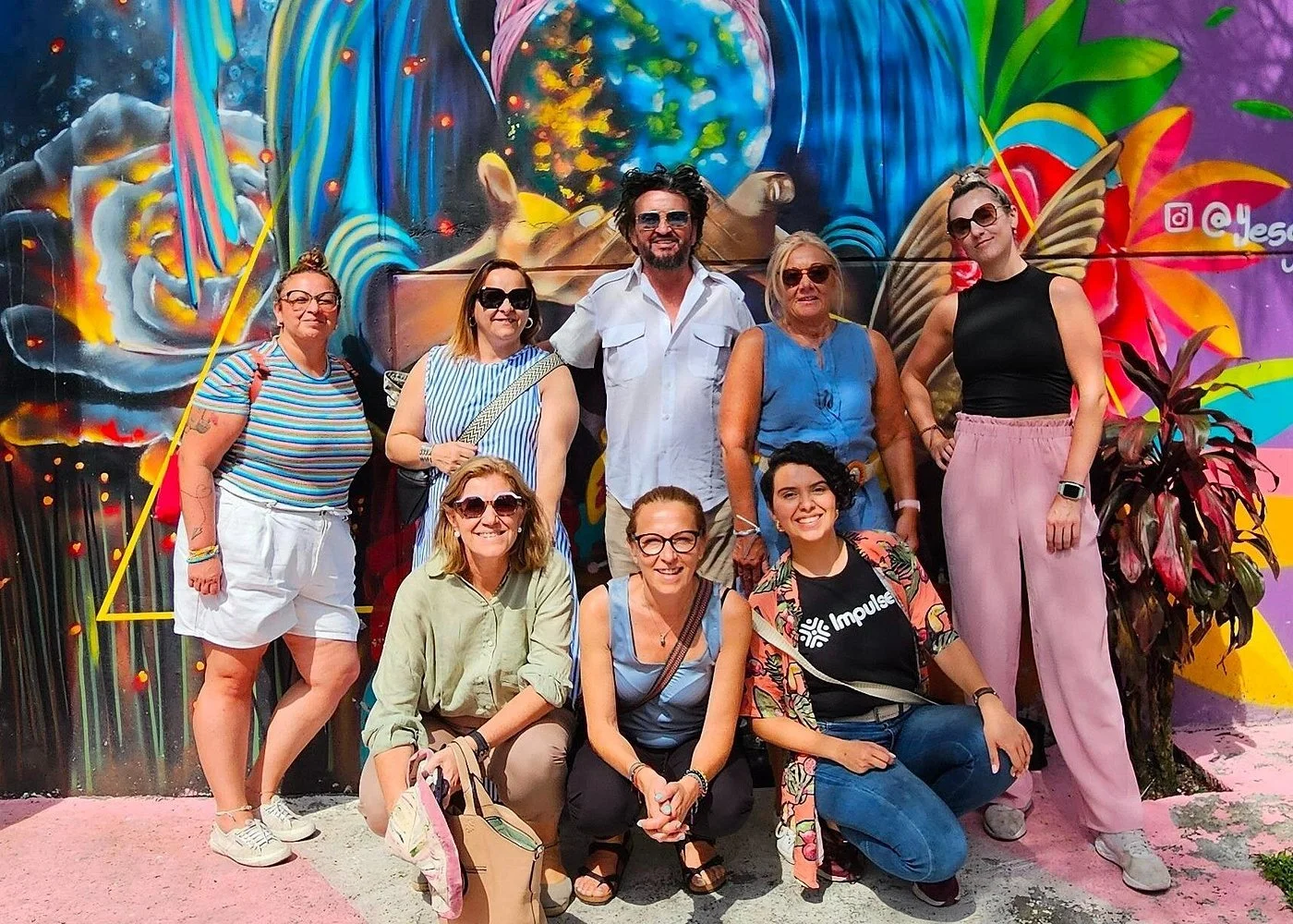 Group of nine women and one man posing in front of a vibrant, colorful graffiti mural featuring flowers, butterflies, and abstract designs.