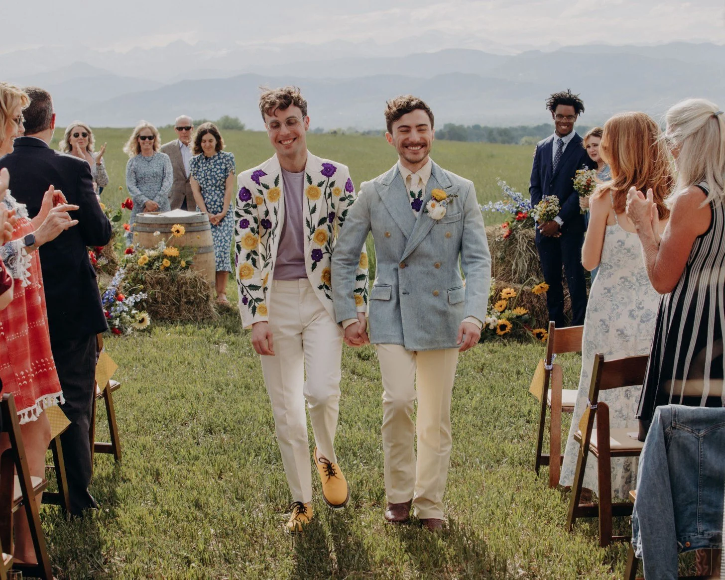 Two men holding hands at an outdoor wedding ceremony with guests clapping and smiling, set in a field with mountains in the background.