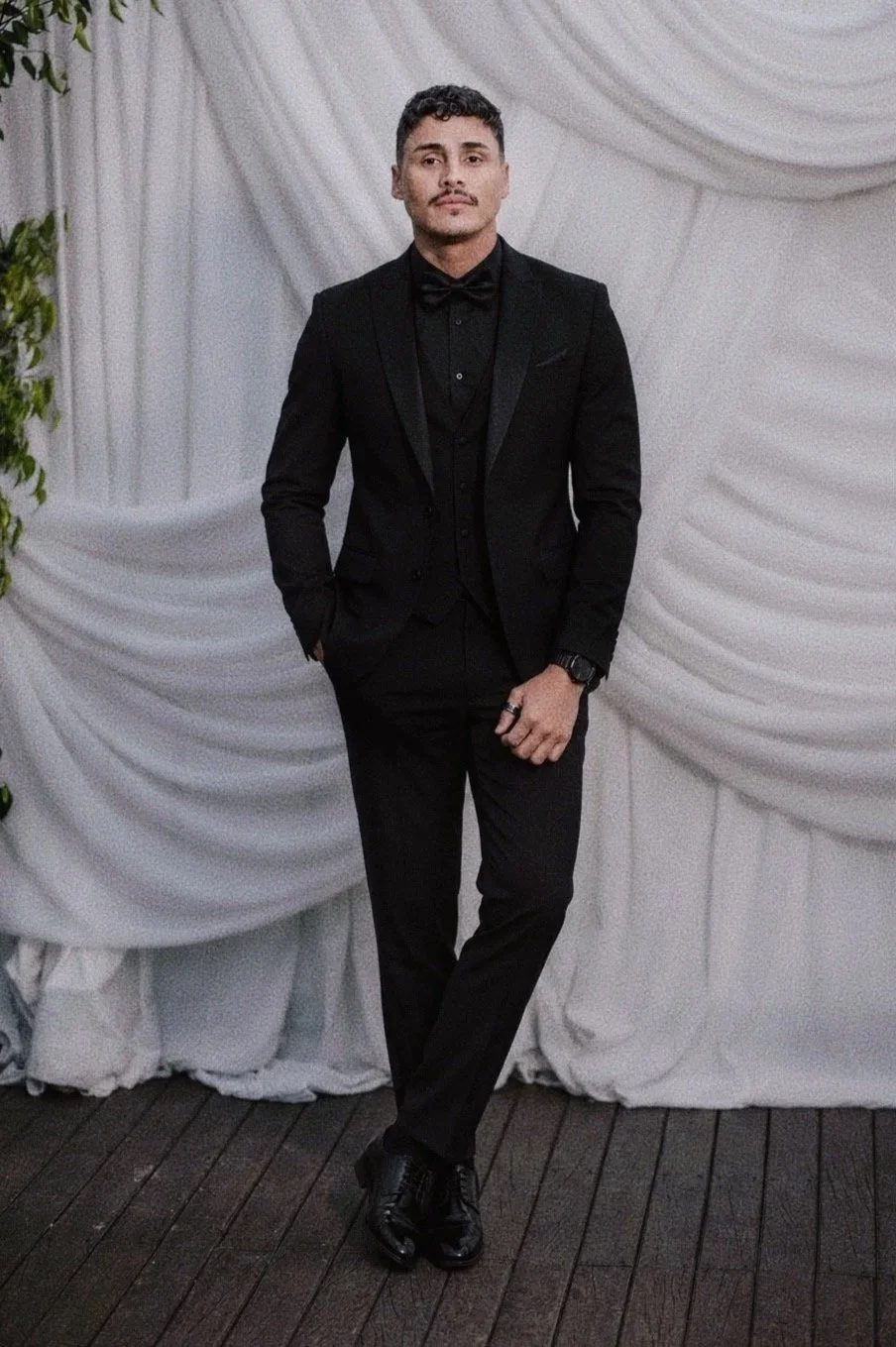 A man dressed in a black tuxedo with a bow tie, standing in front of a white draped curtain background. He has short dark hair, a mustache, and is wearing black shoes and a wristwatch.