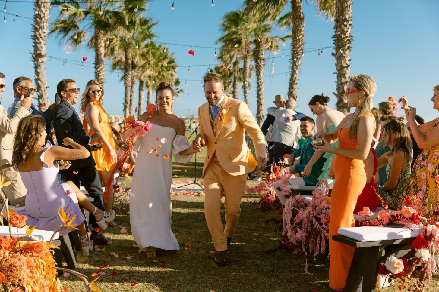 A wedding celebration outdoors with a bride and groom walking down the aisle, surrounded by guests, palm trees, and string lights, with flower petals in the air.