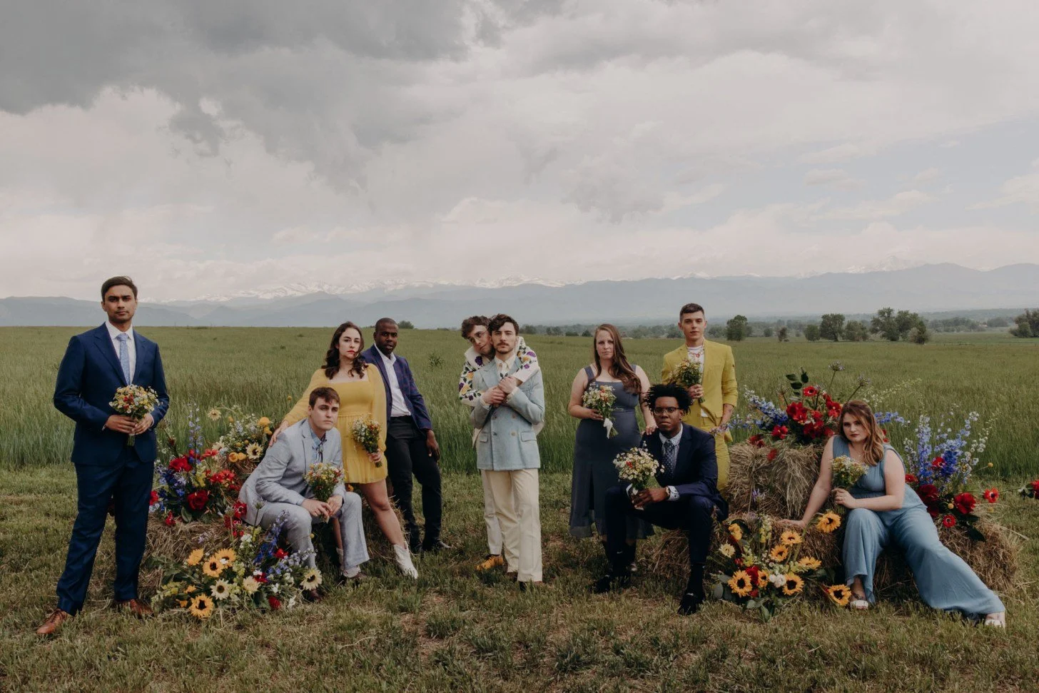 A group of ten people standing and sitting in a field with hay bales, flowers, and a mountain range in the background, dressed in colorful formal and semi-formal attire, under a cloudy sky.
