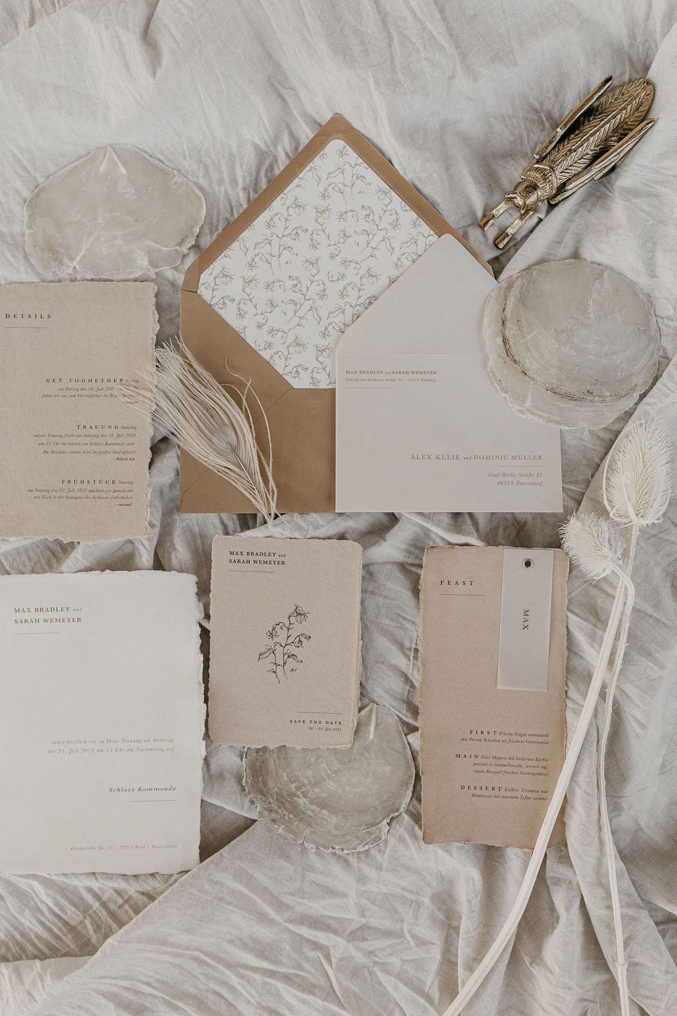 Wedding invitation cards and decorative items on a crinkled, light-colored fabric background, including a carved wooden insect sculpture and textured circular objects.
