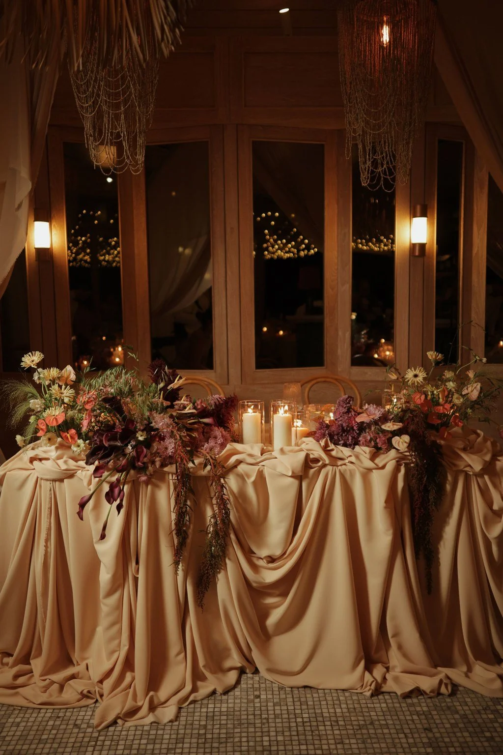 A decorated table with beige draped fabric, floral arrangements with various flowers and greenery, and candles set near a wooden window with curtains, in a warmly lit indoor venue.