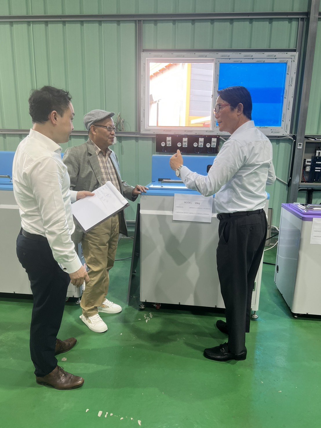 Three men in a factory or laboratory setting discussing equipment, with one man gesturing and the other two listening attentively.