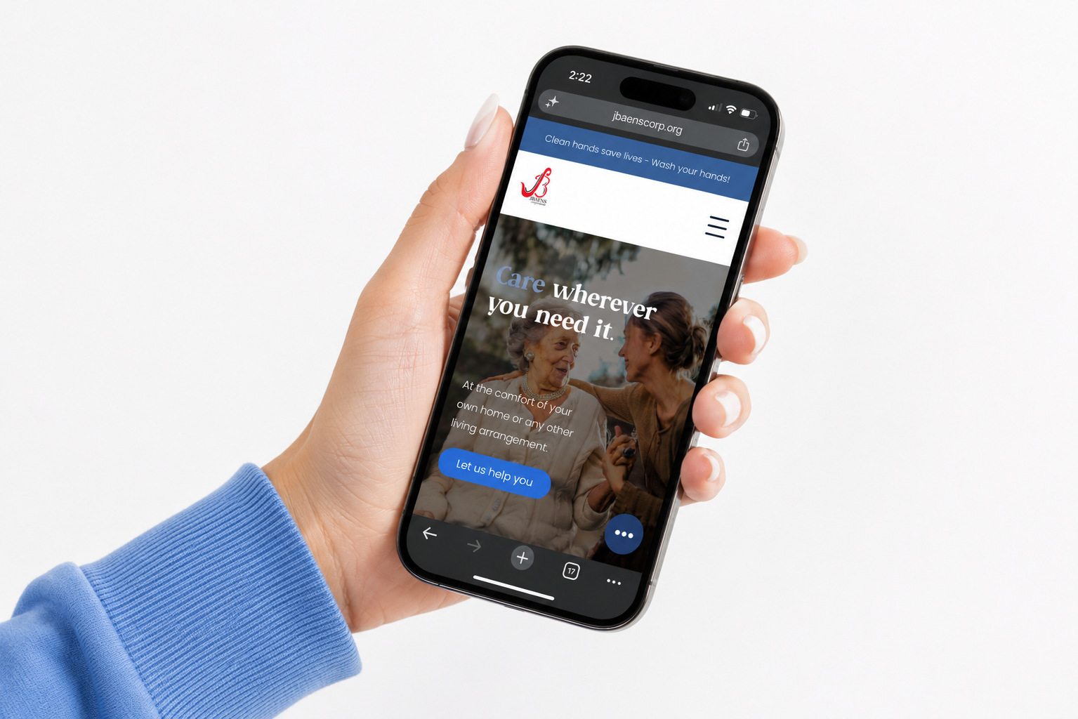 A person wearing a blue long sleeve shirt holding a smartphone with a website for a healthcare or home care organization showing. The website features an elderly woman and a caregiver, with the text 'Care wherever you need it' and a blue button that says 'Let us help you'.