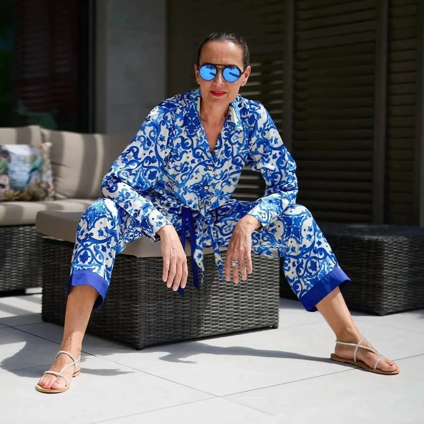 Une femme assise en tailleur sur une banquette en rotin, portant un pyjama bleu et blanc avec un motif floral, des lunettes de soleil bleues et des sandales plates, posée dans un espace extérieur lumineux.