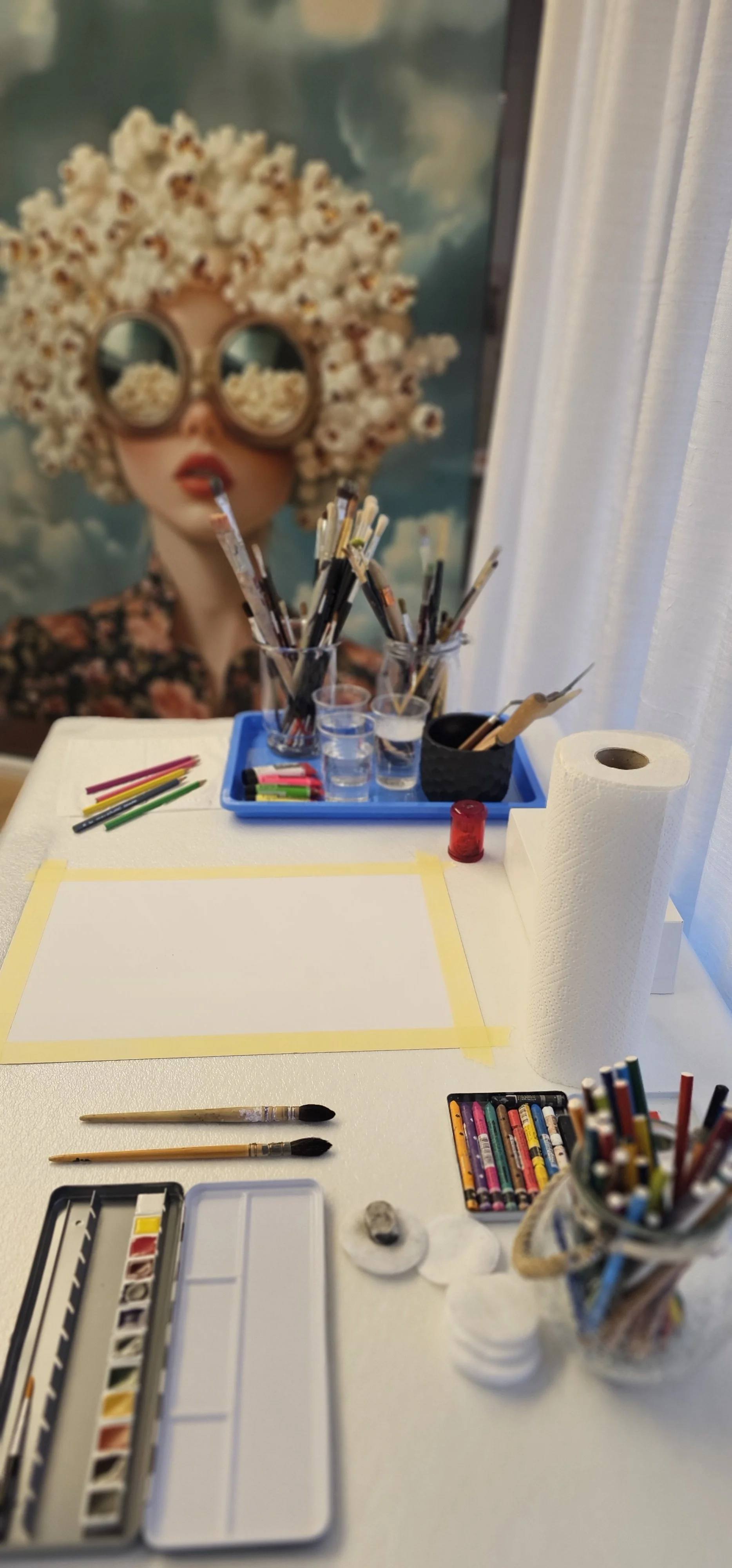 Espace de création artistique avec pinceaux, crayons colorés, papier, rouleau de papier toilette, et une affiche d'une femme avec des lunettes rondes et un chapeau en pop-corn.
