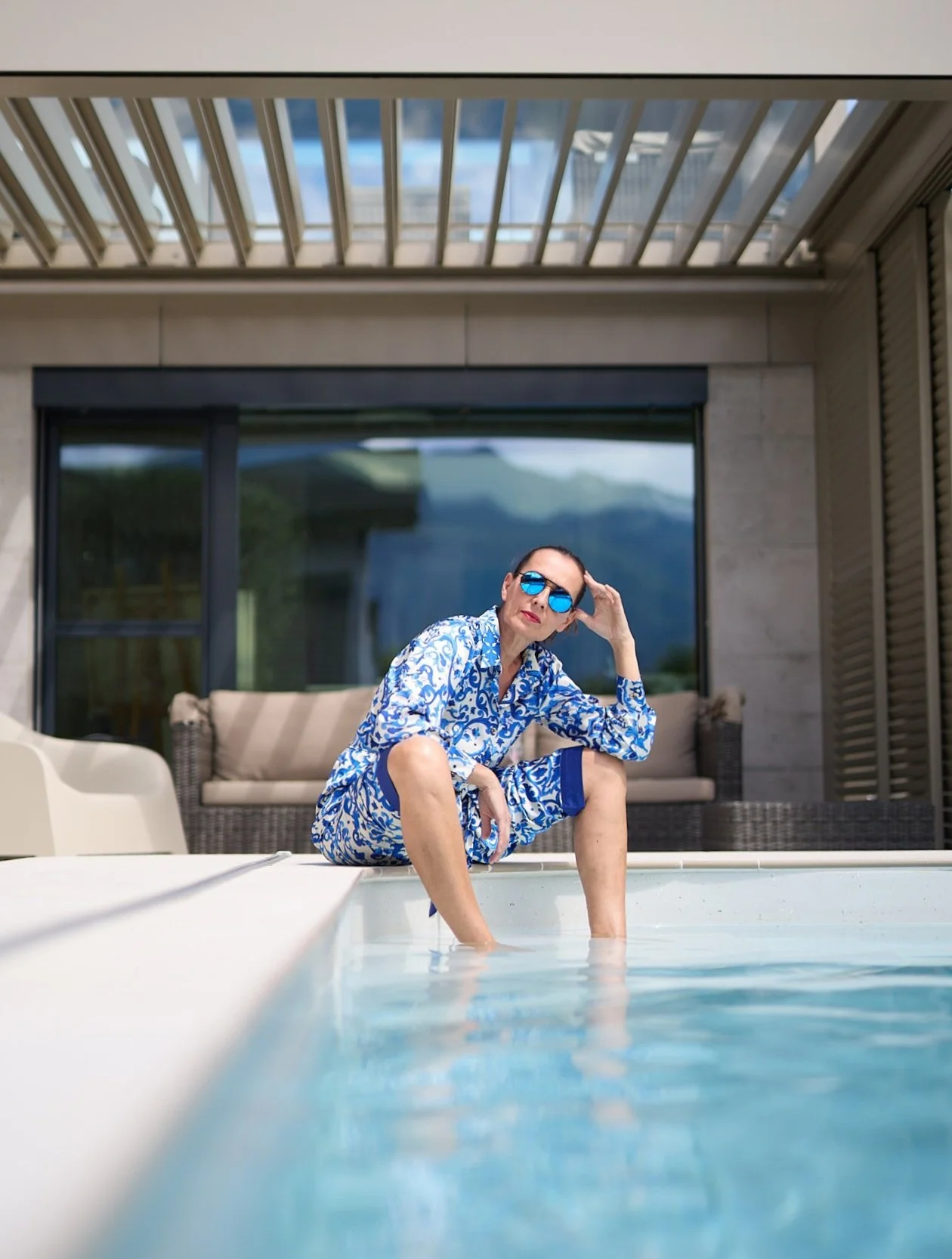 Une femme assise au bord d'une piscine, portant une tenue de plage bleue et blanche avec des lunettes de soleil bleues, devant une maison moderne avec un grand patio et des montagnes en arrière-plan.