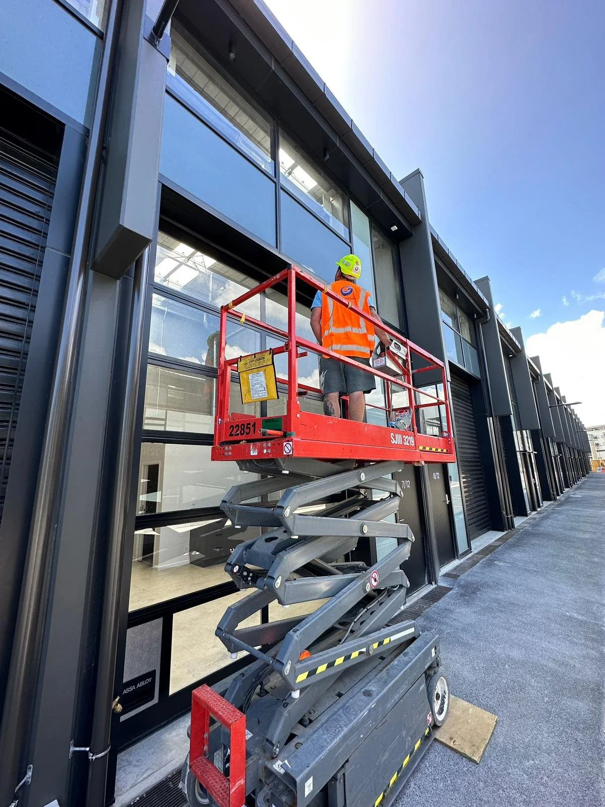 A worker on a red lift platform wearing an orange safety vest and a green helmet with a yellow logo is cleaning a large glass window on the exterior of a modern commercial building with black metal frames, under a blue sky.