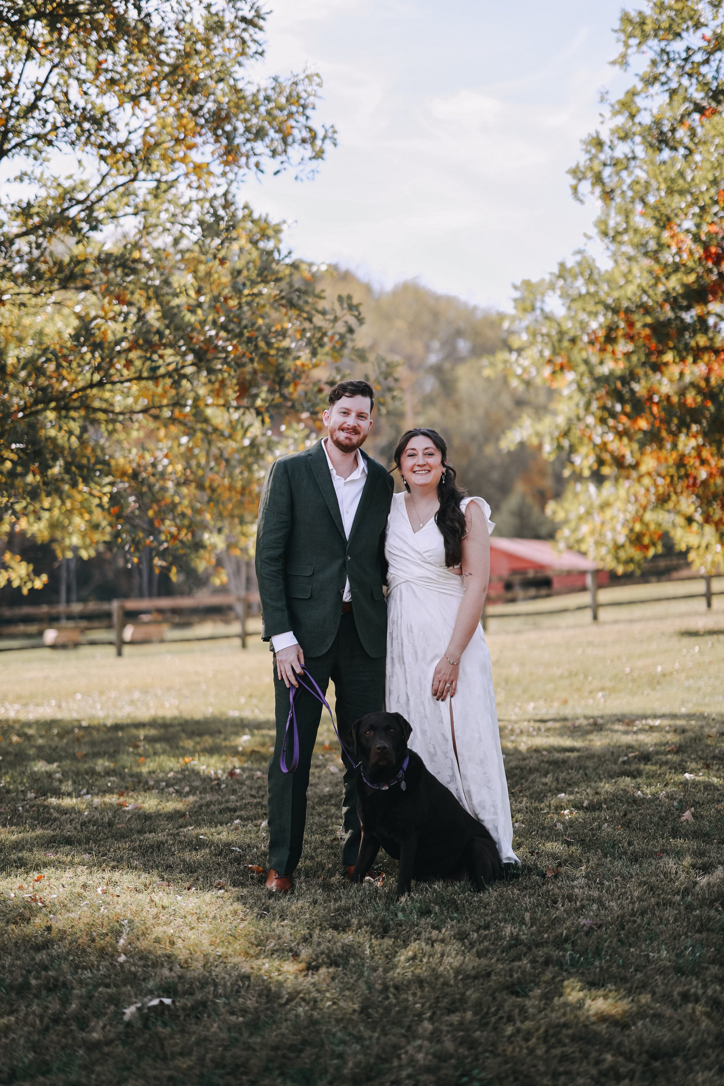 "Jay Hunter and his wife Hannah stand with their chocolate Labrador retriever Luka at their wedding reception in a Virginia autumn landscape, with a red barn and fall foliage in the background."