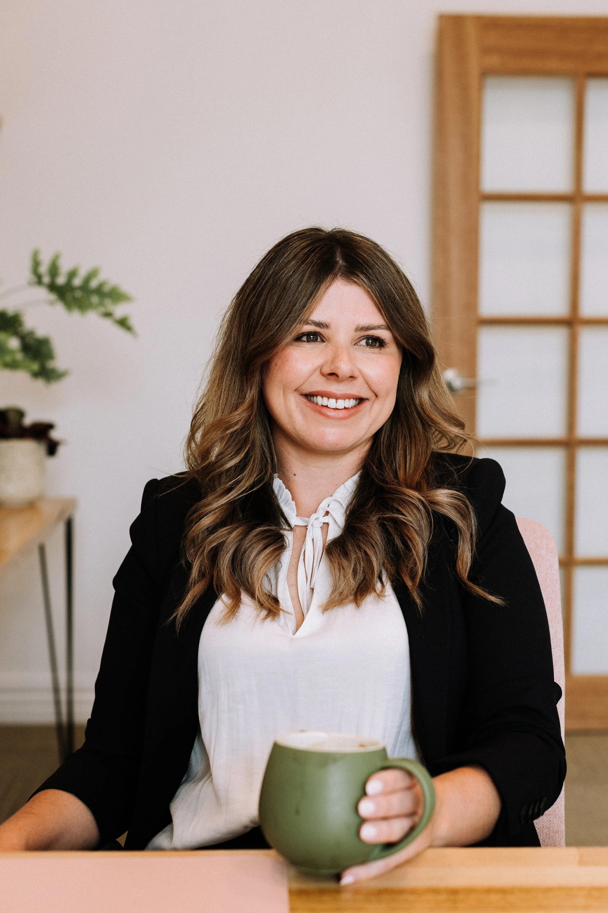 A woman with long brown hair smiling and holding a green mug, sitting at a table in a bright room with a wooden door and a plant in the background.