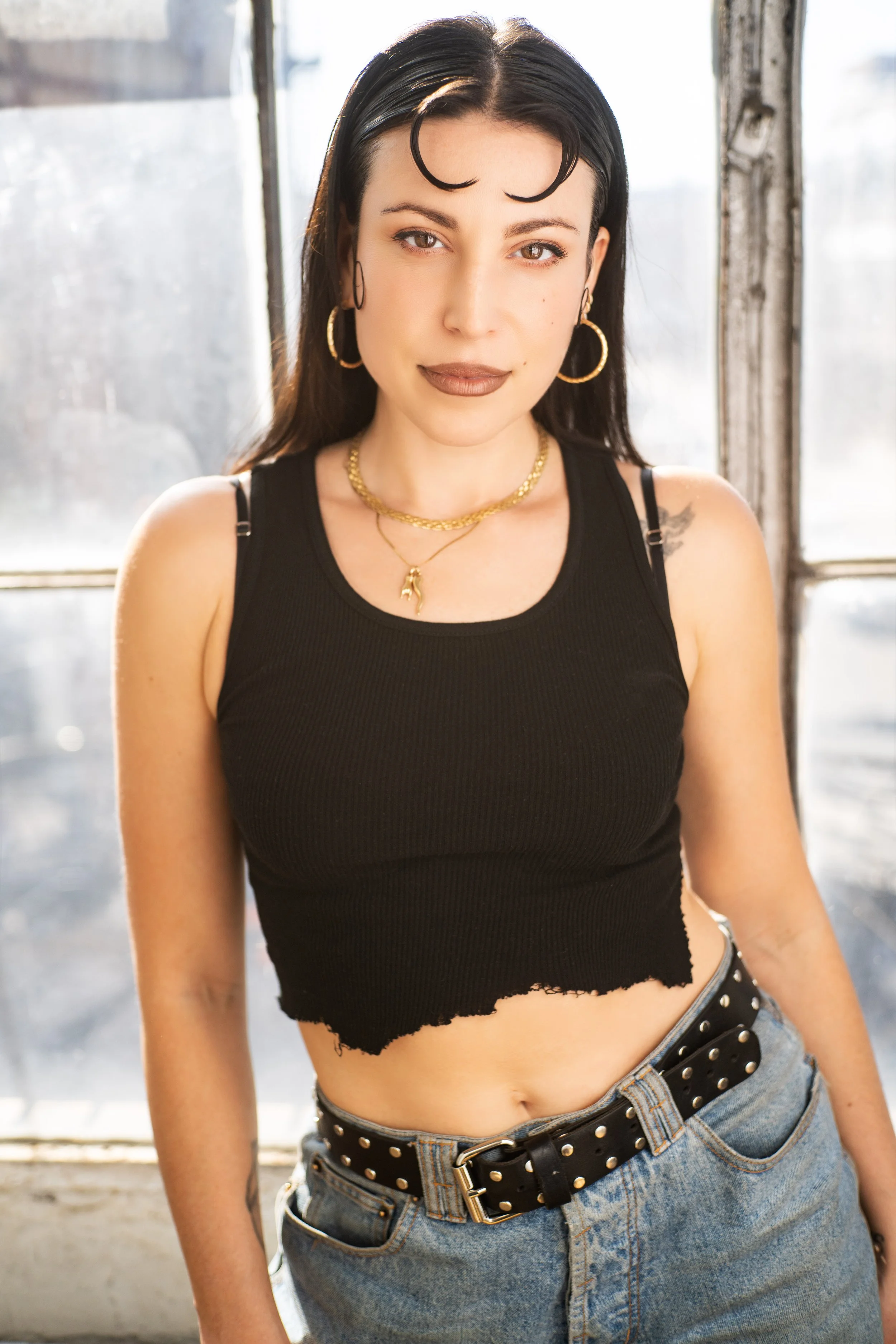A young woman with dark hair and hoop earrings stands in front of a window, wearing a black sleeveless top and denim pants with a black studded belt.