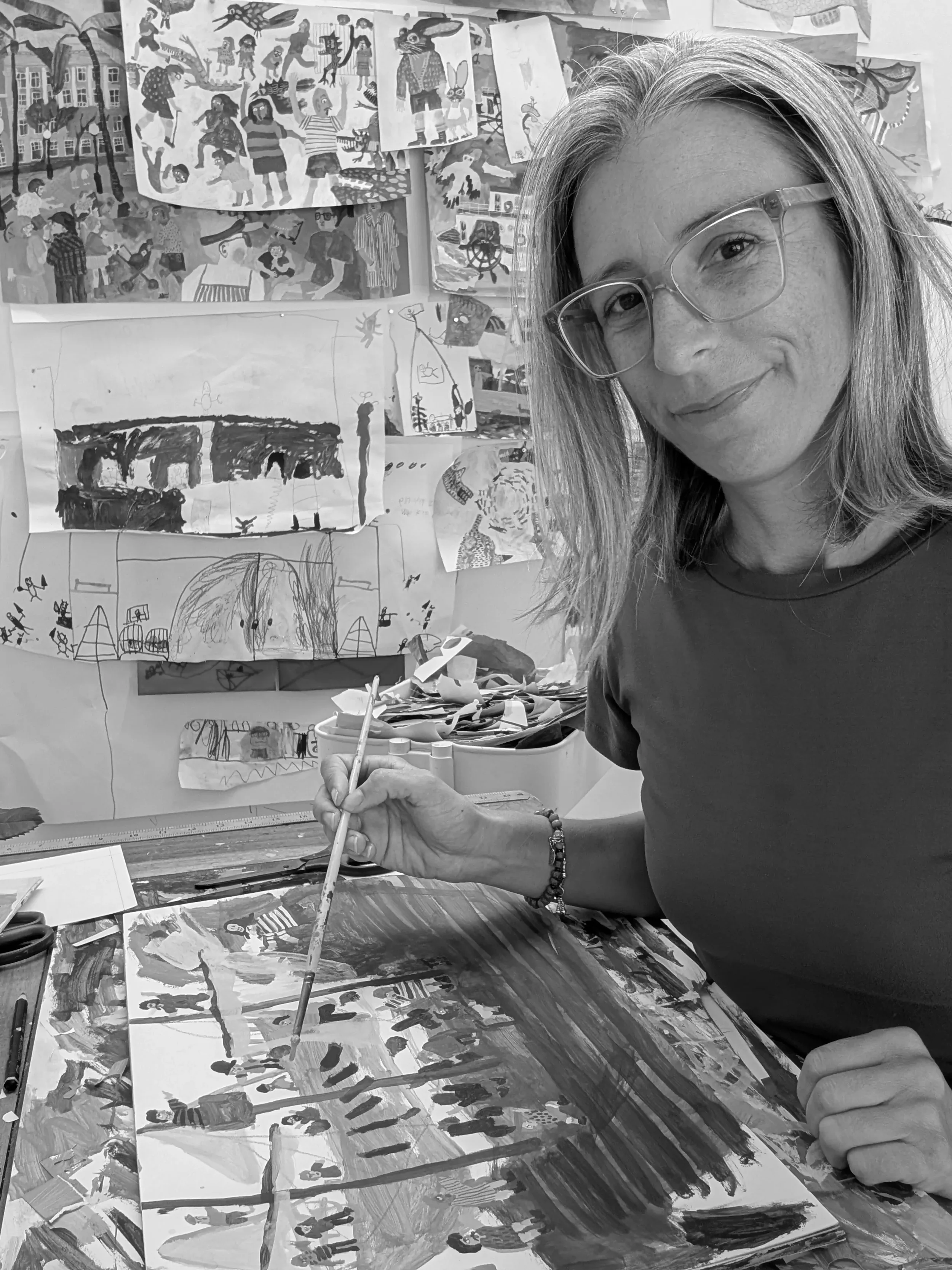 A woman wearing glasses and a dark shirt, smiling while painting a large colorful artwork on a table. Behind her is a wall covered with many smaller colorful drawings and paintings, including various figures, animals, and scenes.