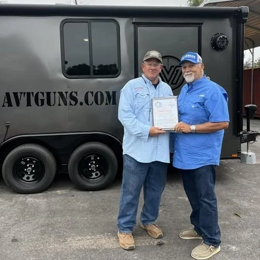 Two men stand outside in front of a black trailer with a window and the website AVTGUNS.COM printed on it. They are holding a document together, smiling at the camera. One man wears a light blue shirt and beige boots, and the other wears a blue shirt with a blue cap and gray shoes.